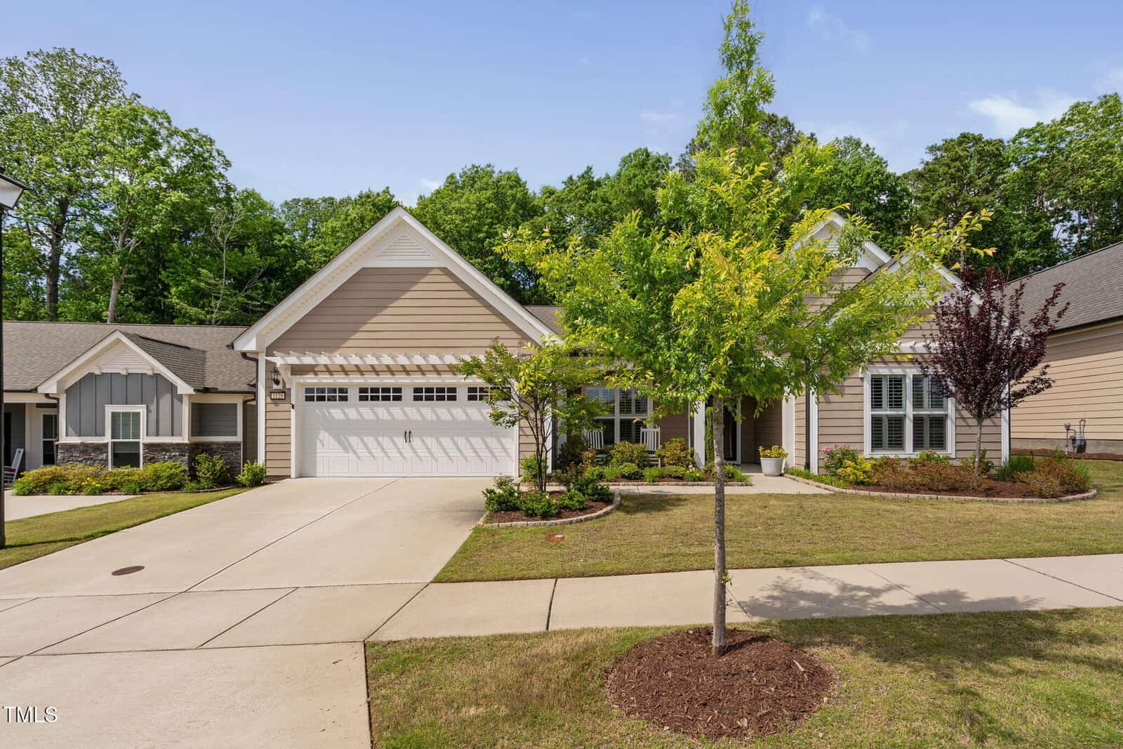 Single-story suburban house with a double garage, beige siding, and white trim. Manicured lawn, young trees, and landscaped flower beds in front. Surrounded by greenery and adjacent houses.