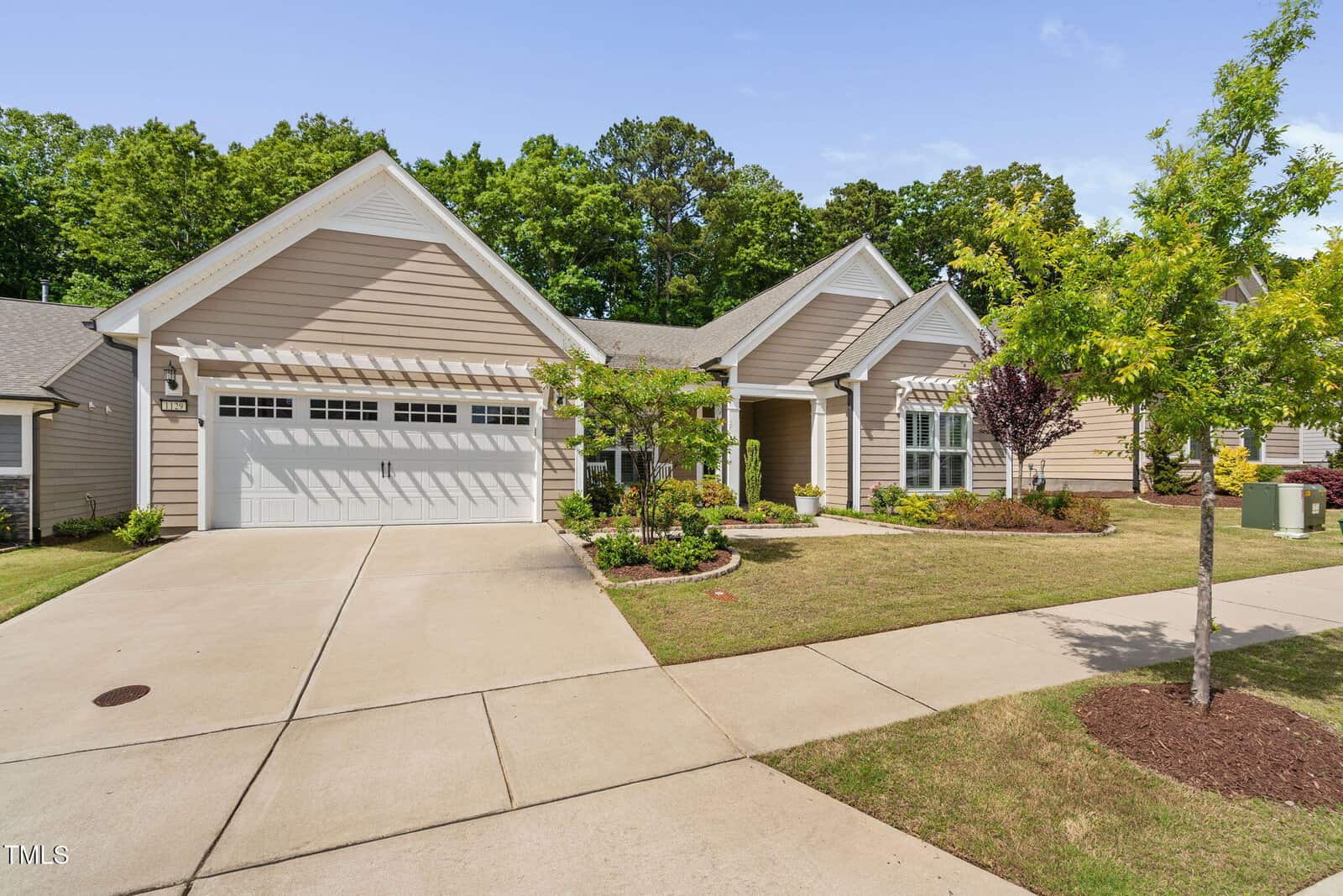 Single-story beige house with white trim, double garage, and well-maintained front yard featuring small trees, green grass, and shrubs. Concrete driveway leads to the garage. Trees and neighboring houses are visible in the background.