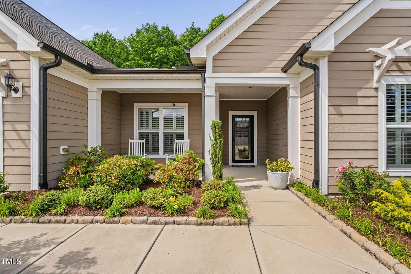 A beige house with white trim features a covered front porch, two white rocking chairs, landscaped bushes, flowers, and a walkway leading to a glass front door.