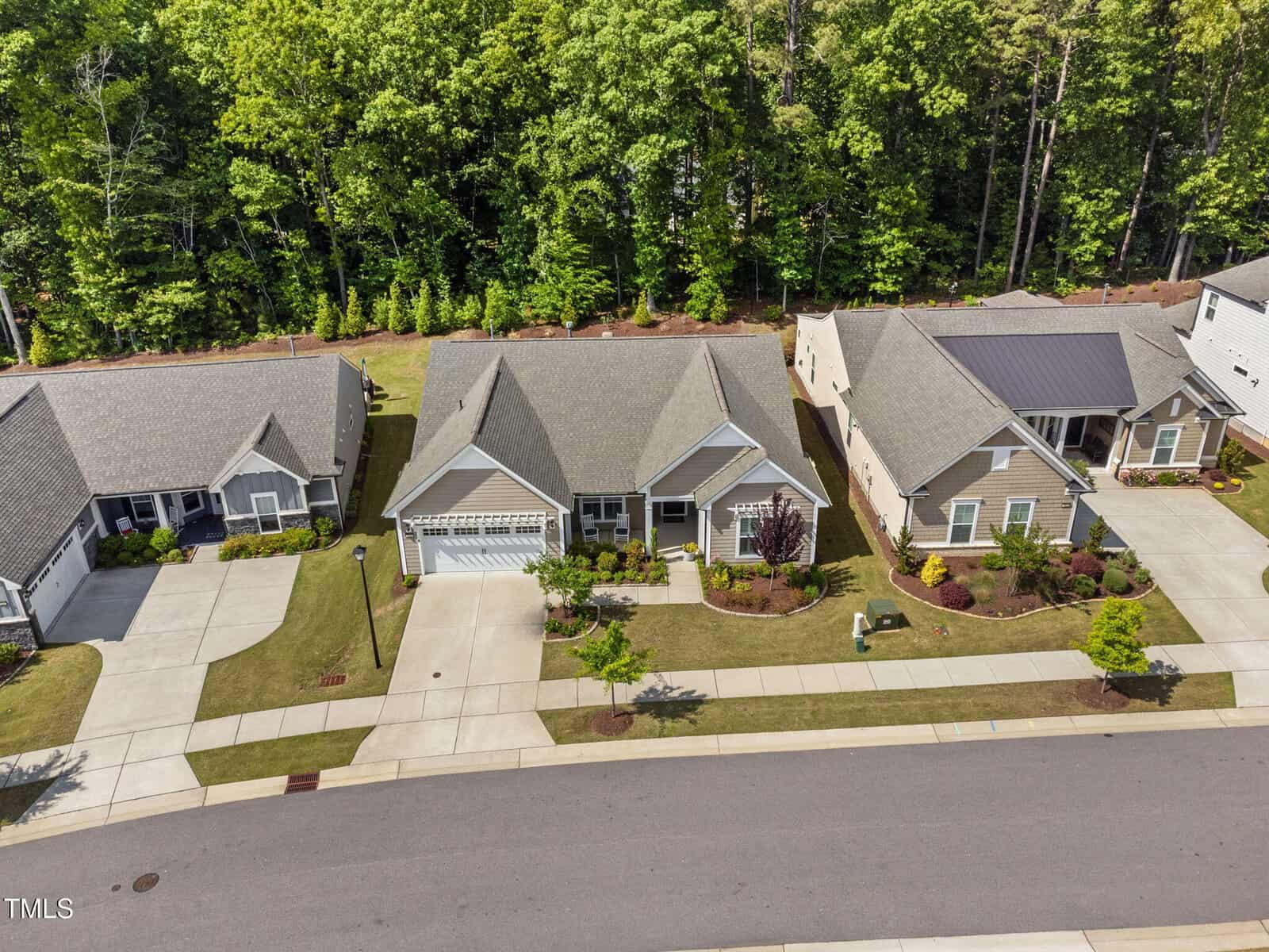 Aerial view of three single-story suburban houses with neatly manicured lawns, driveways, and sidewalks. Behind the houses is a dense row of tall green trees. The street and part of the road are visible in the foreground.