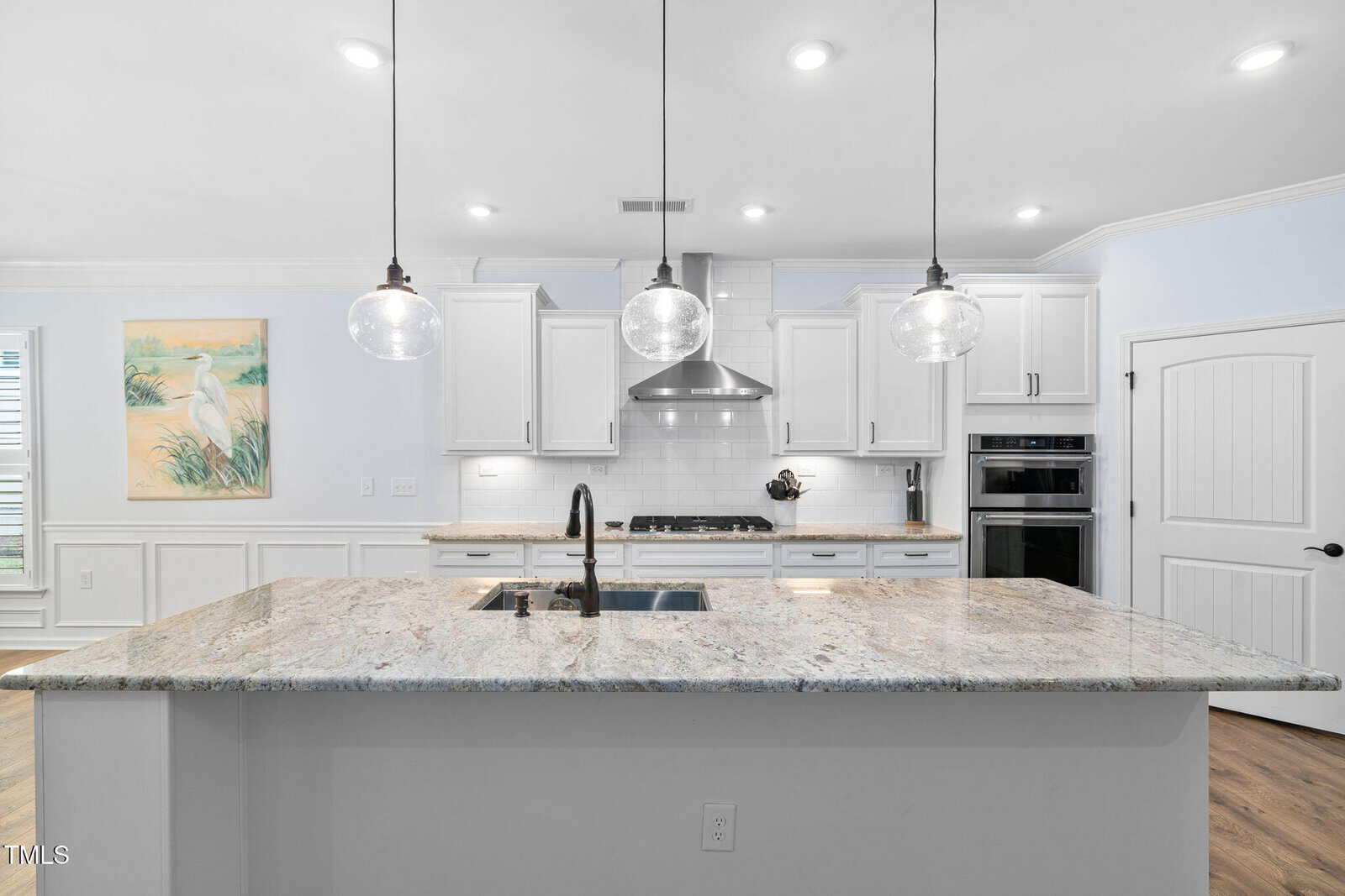 Modern kitchen with a large granite island, stainless steel sink and appliances, white cabinets, pendant lights, a range hood, built-in oven, and light wood flooring; a painting of a bird hangs on the left wall.