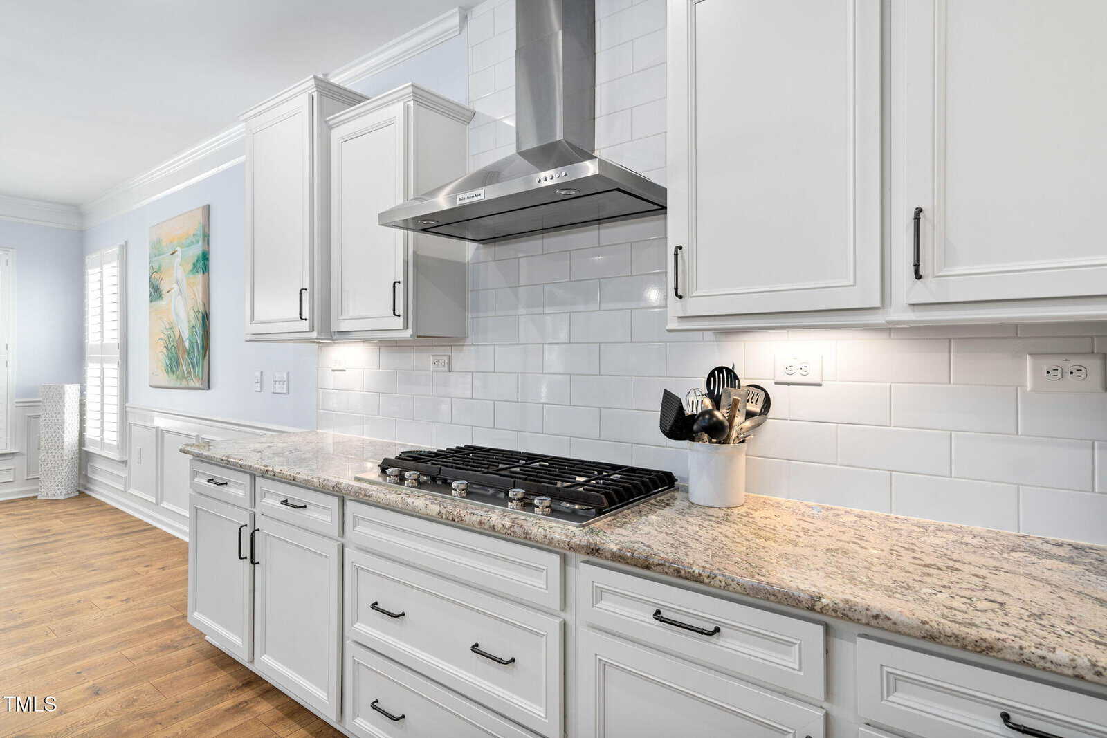 Modern kitchen with white cabinets, granite countertop, stainless steel gas cooktop and hood, white subway tile backsplash, and a utensil holder. Under-cabinet lighting brightens the workspace. Artwork hangs on the wall to the left.