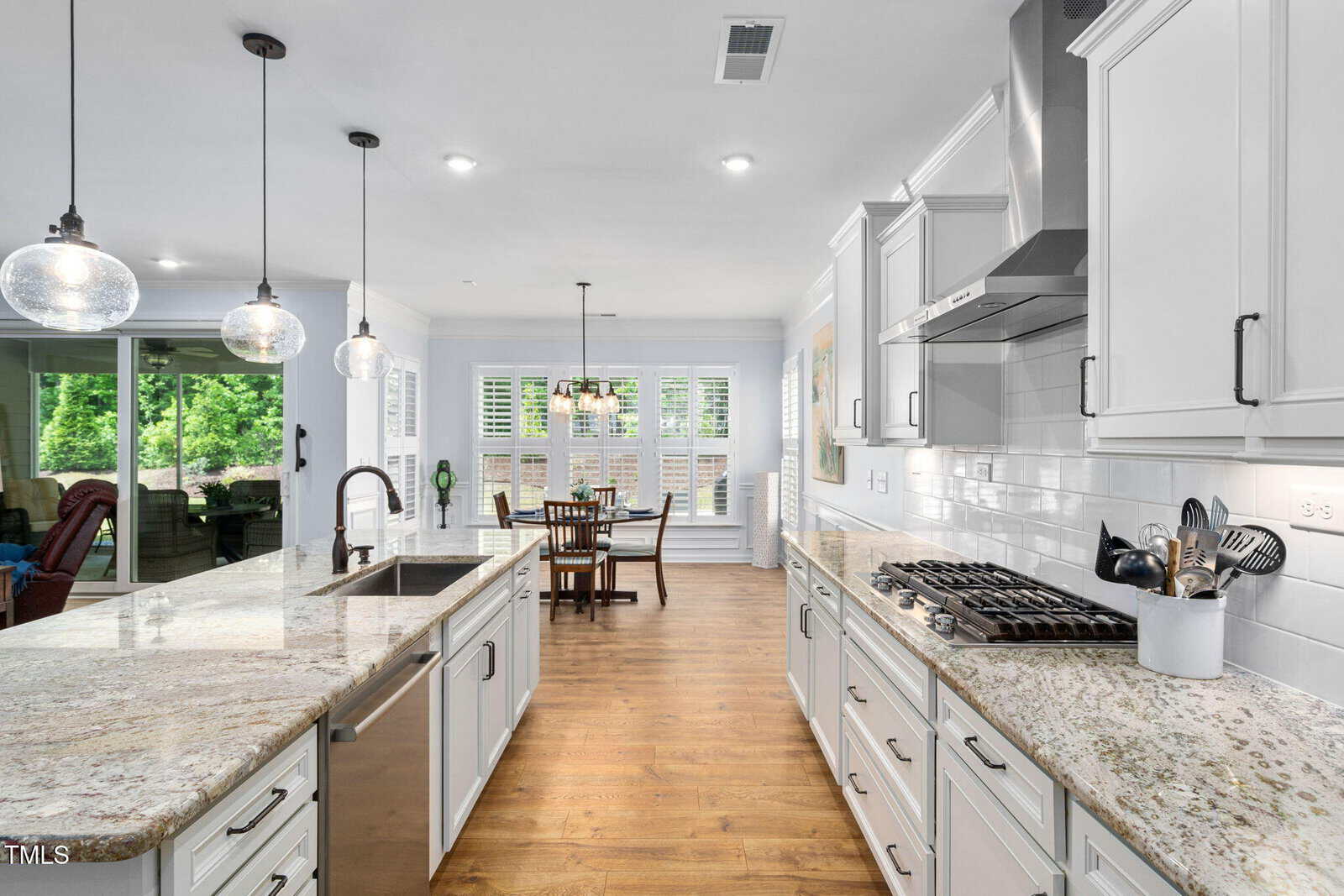 Bright, modern kitchen with white cabinets, granite countertops, stainless steel appliances, and a gas cooktop. An island with a sink faces an open dining area with large windows. Hardwood floors throughout.