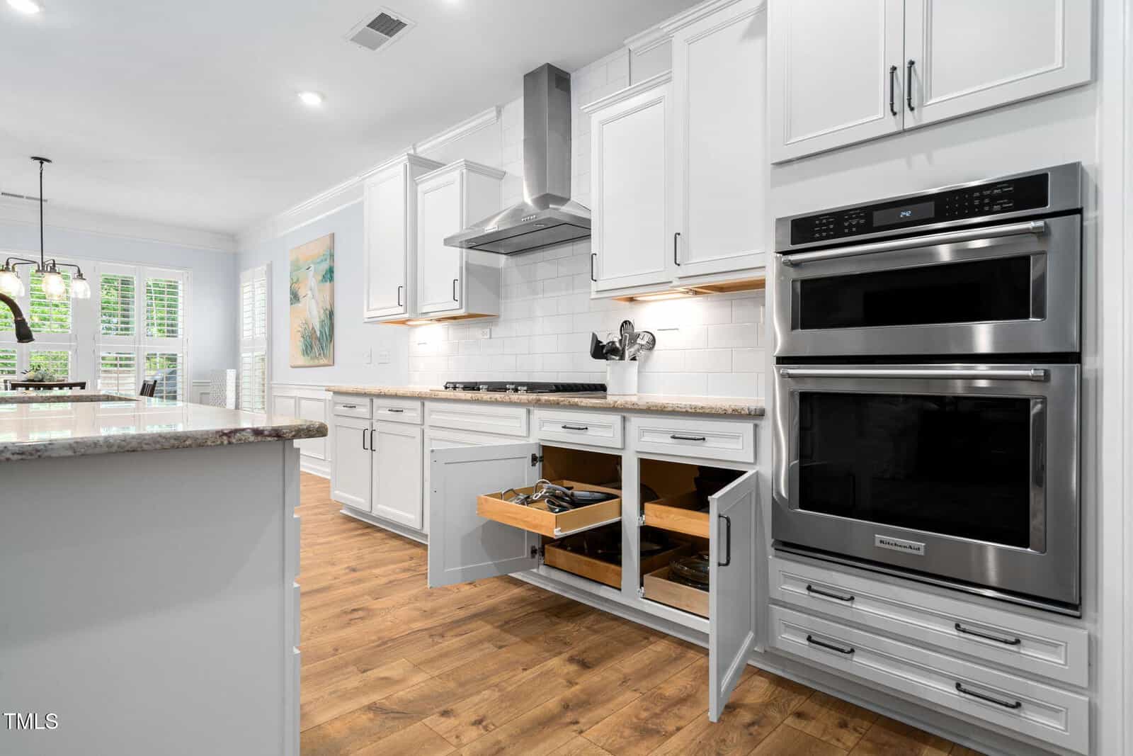 Modern kitchen with white cabinets, stainless steel appliances, granite countertops, and wood flooring. Two lower cabinet drawers and one cabinet door are open, revealing storage. Dining area and large windows are visible in the background.