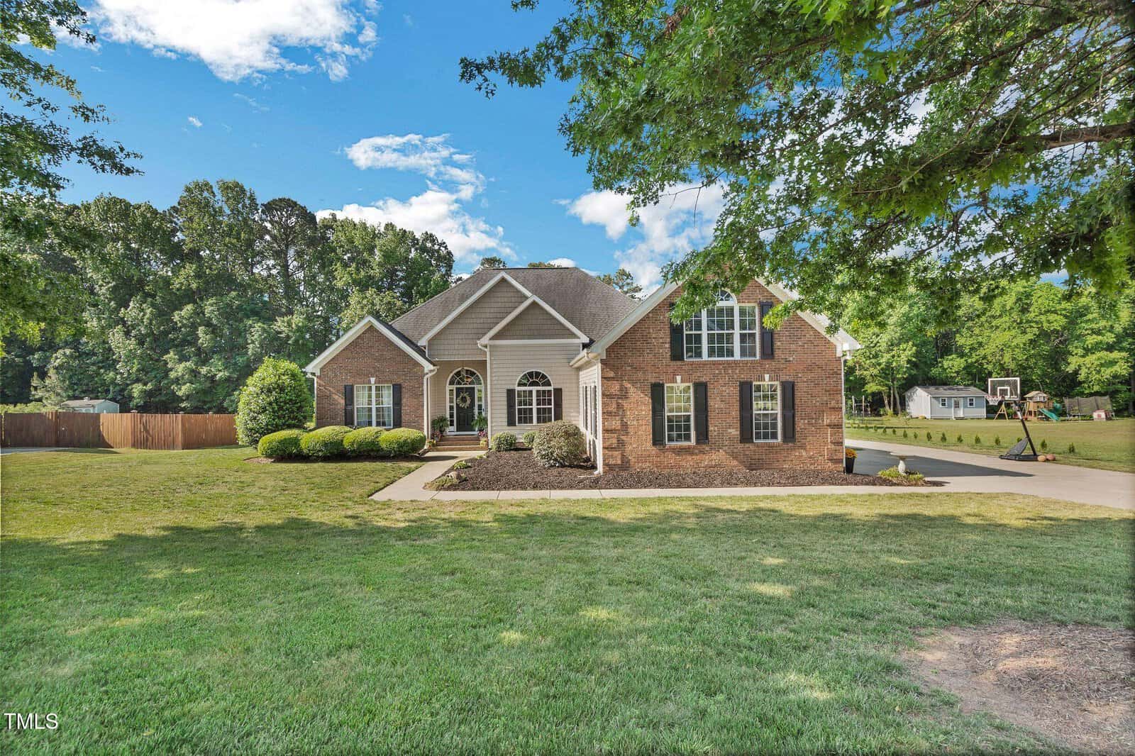 Single-story brick and siding house with a well-manicured lawn, shrubs, arched front porch, and a long driveway, set against a backdrop of trees and a partly cloudy blue sky.