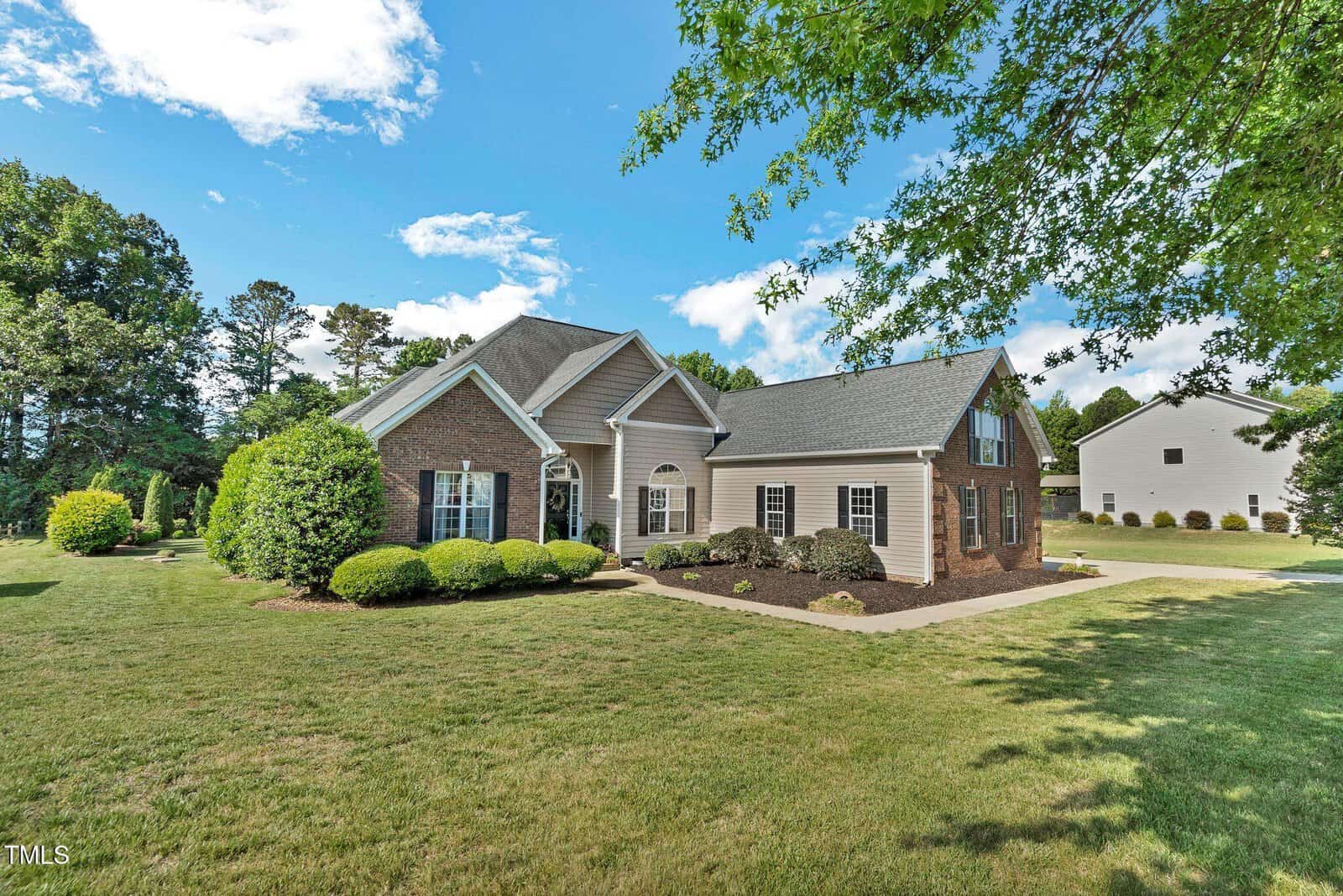 A single-story brick house with white trim, gabled roof, and multiple windows sits on a large, well-maintained lawn surrounded by trees and shrubs under a partly cloudy blue sky.