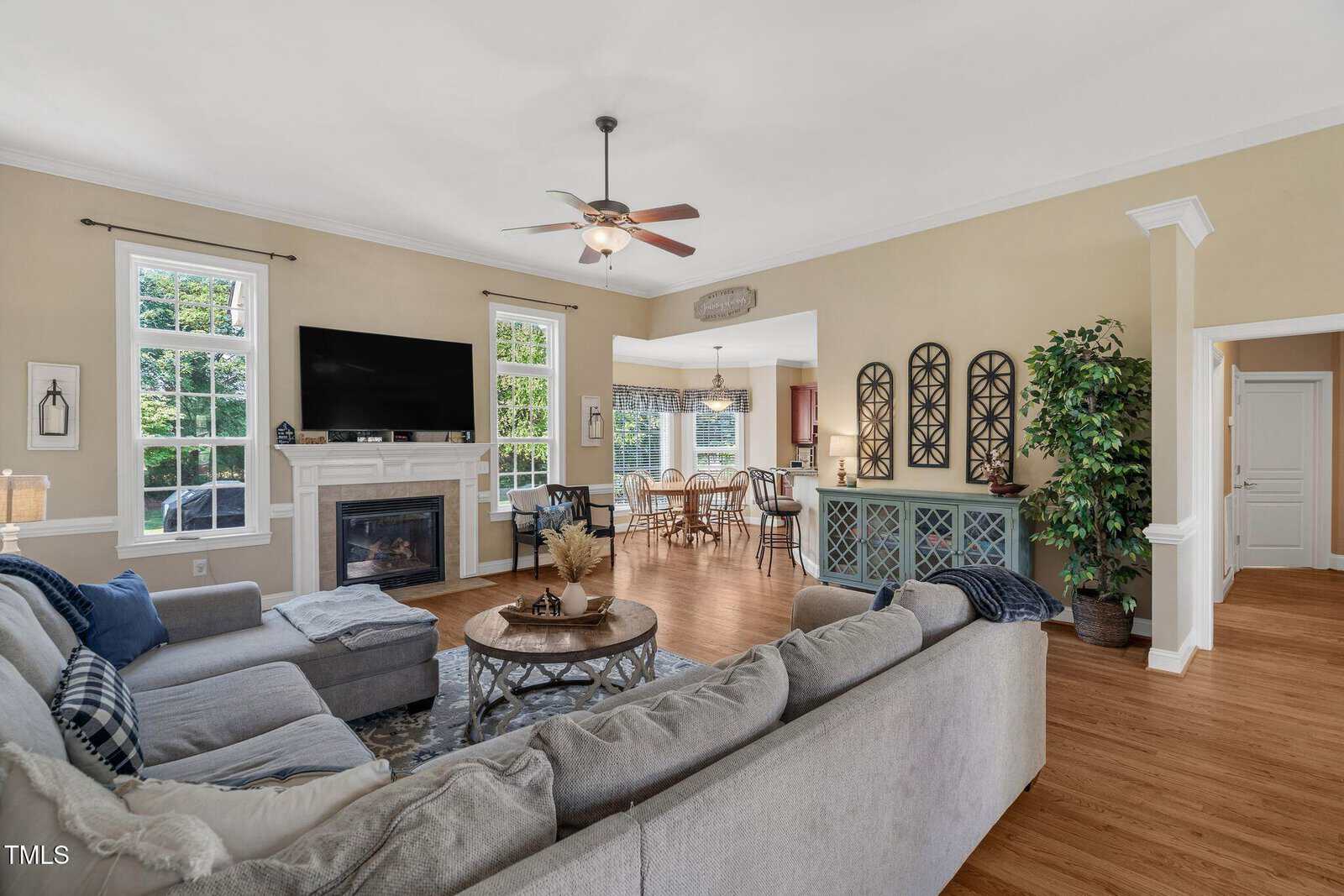 A spacious living room with a gray sectional sofa, wall-mounted TV above a fireplace, hardwood floors, large windows, and a view into a dining area with a wooden table and chairs. Decor includes wall art and indoor plants.