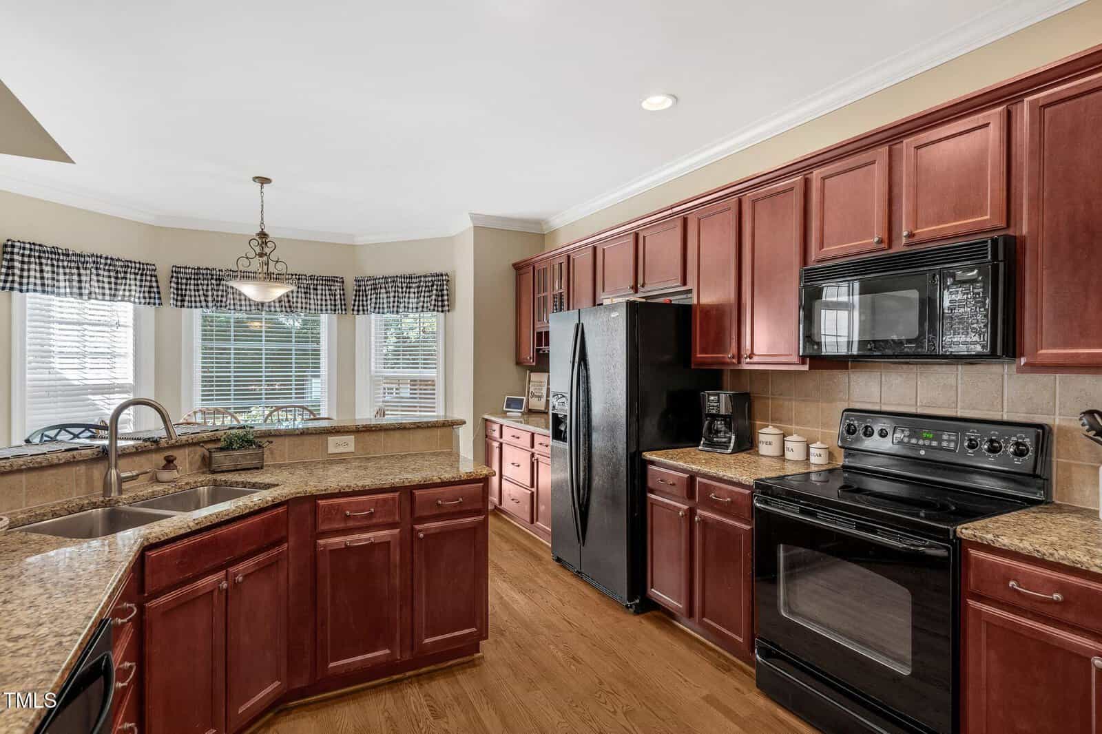 Spacious kitchen with dark wood cabinets, granite countertops, black appliances, tile backsplash, double sink under large windows with plaid valances, and hardwood floors. A chandelier hangs above the dining area.