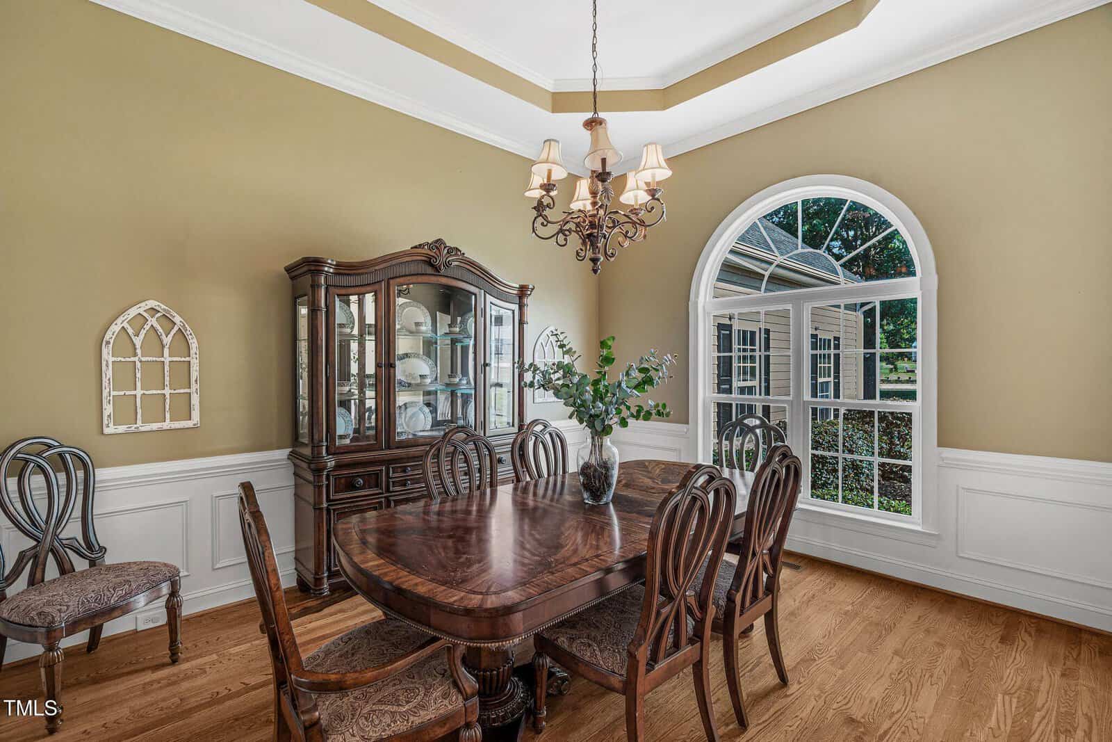 Formal dining room with a wooden table and six chairs, a china cabinet, hardwood floors, beige walls with white wainscoting, a chandelier, arched window, and decorative wall accents. Greenery visible outside the window.