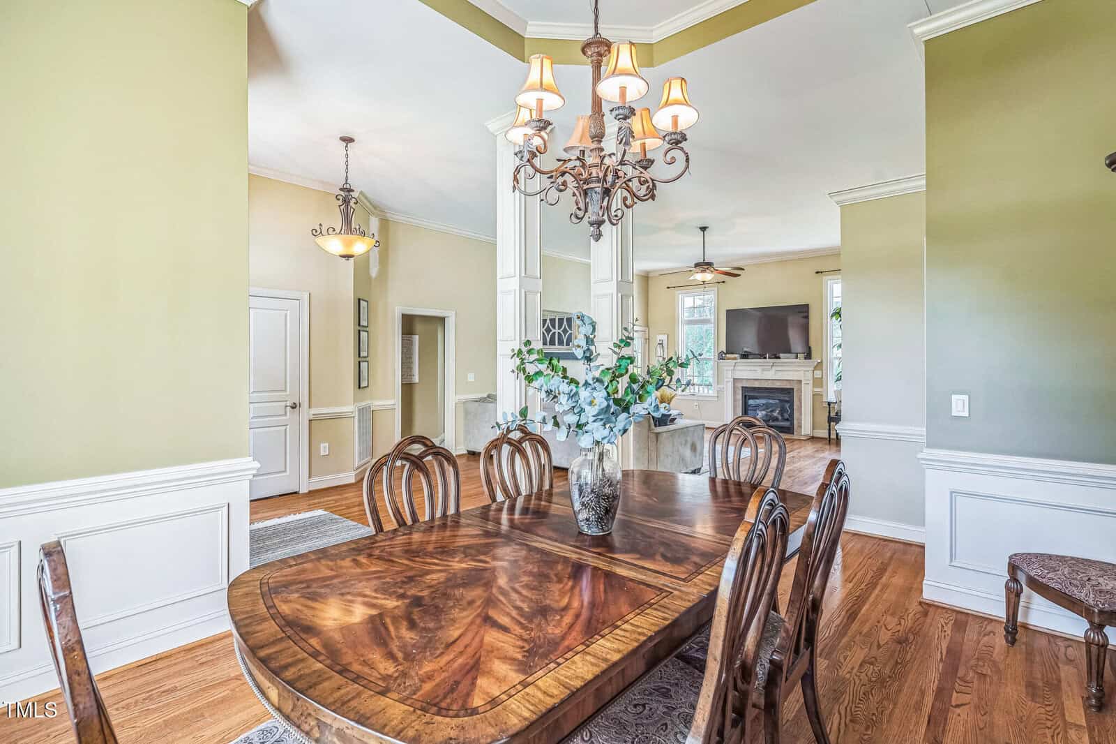 Dining room with a wooden table and chairs, a chandelier overhead, light green walls with white trim, hardwood floors, and a vase with white flowers on the table. A view into a living room and hallway is visible.