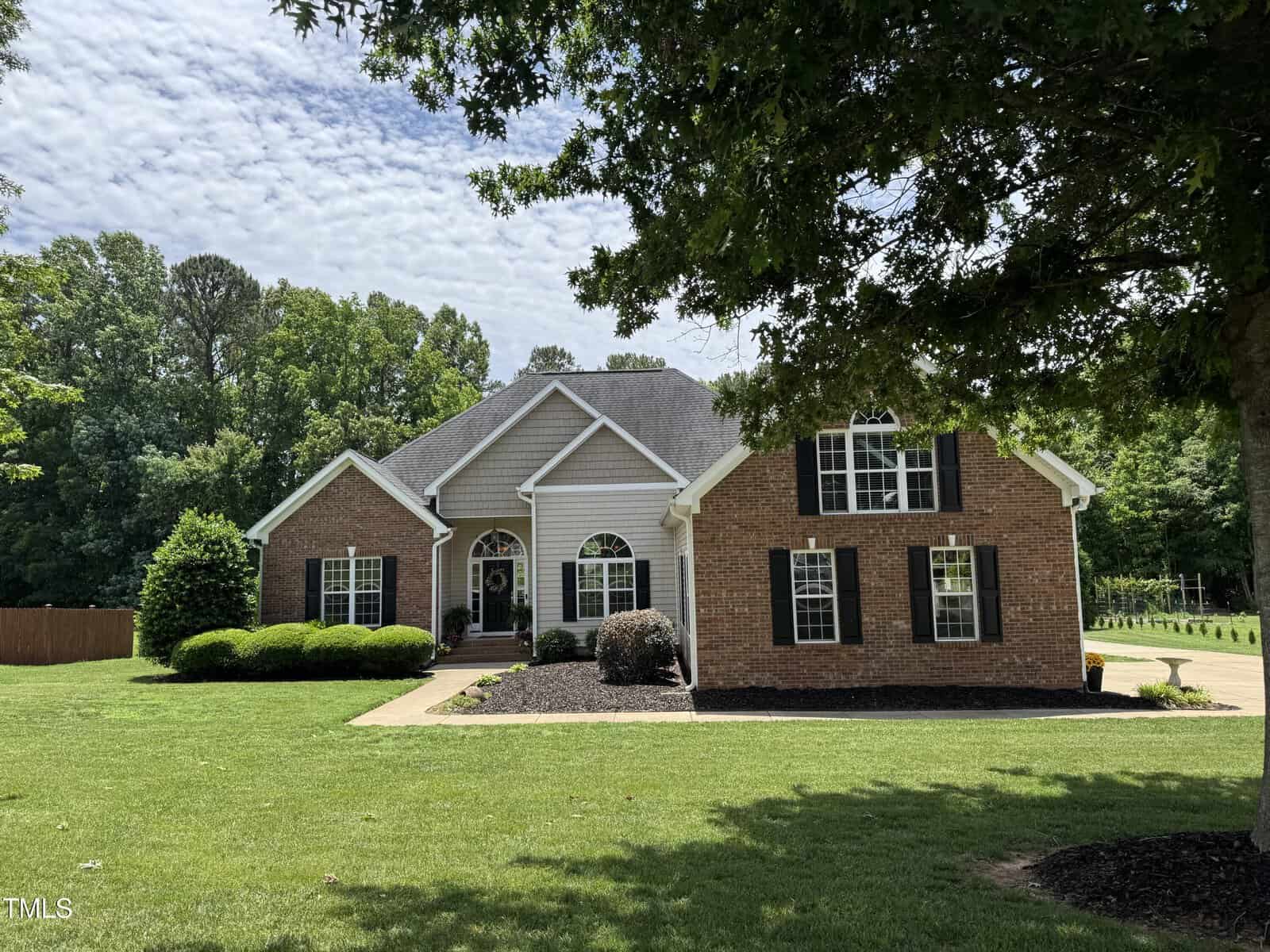 A one-story suburban brick house with white trim, large windows, and an arched entryway sits on a well-kept lawn. Trees and shrubs surround the house, and a driveway leads up to the front.