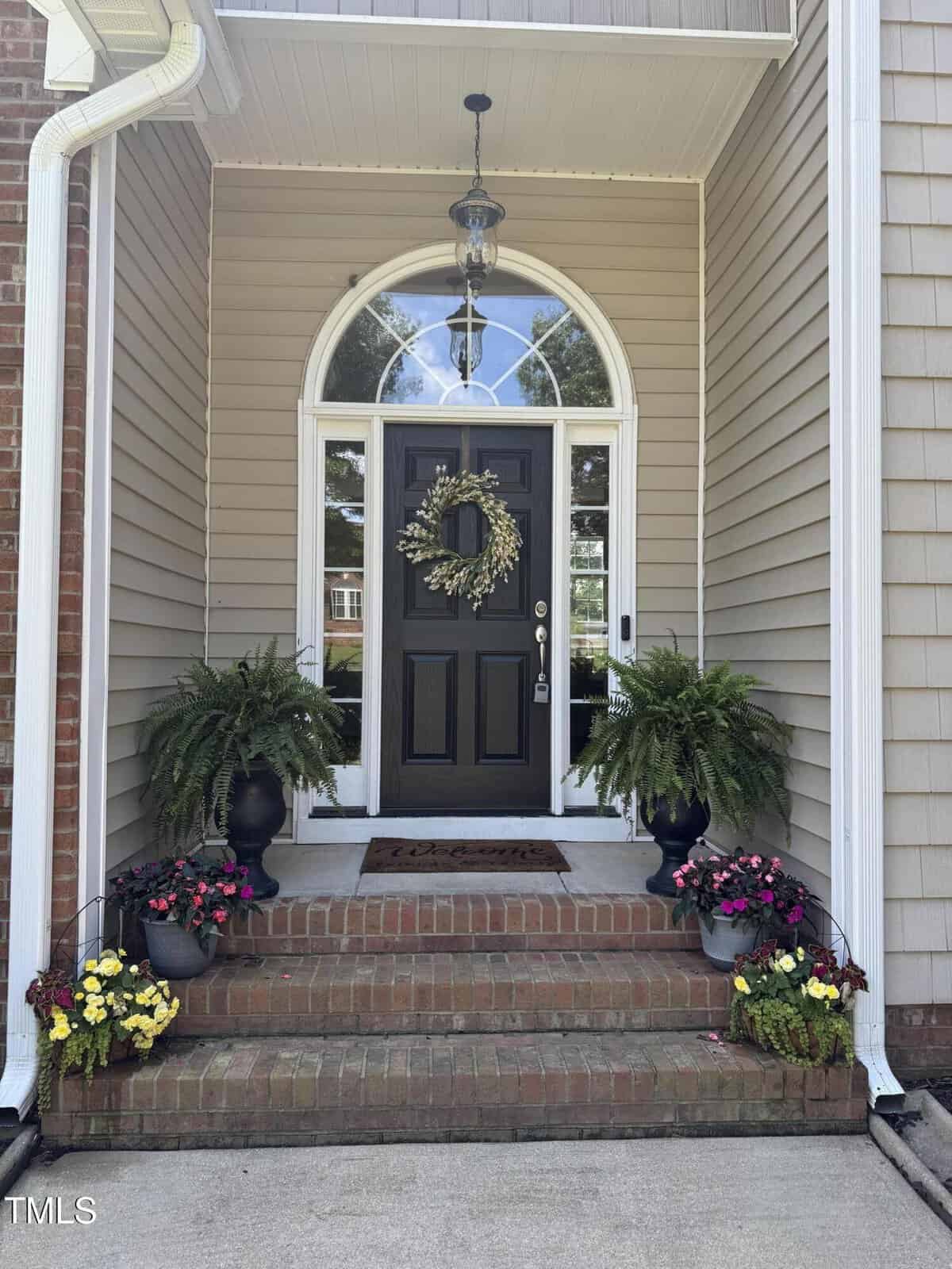 A black front door with a wreath, set in a white frame and arched window, is flanked by two potted ferns. Brick steps lead up, lined with colorful flowers in pots on each side. Beige siding and a hanging light are visible.