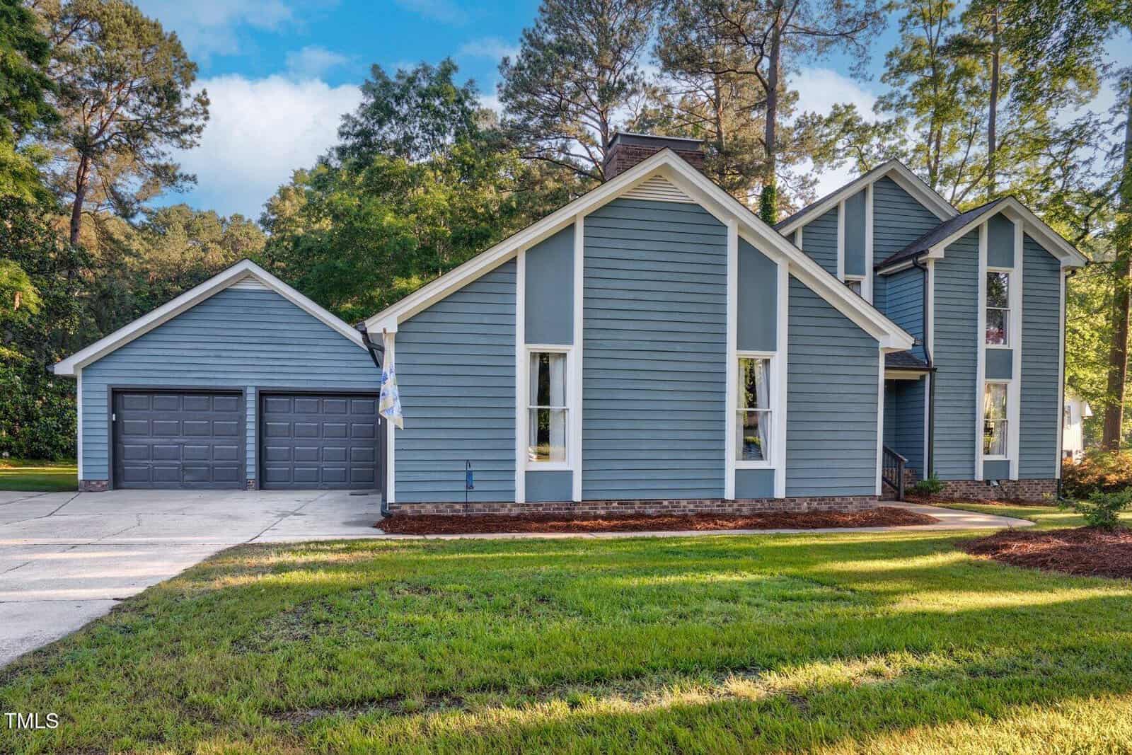 A two-story blue house with white trim and a brick foundation, attached two-car garage with gray doors, and a concrete driveway. The yard has green grass, and there are tall trees in the background.
