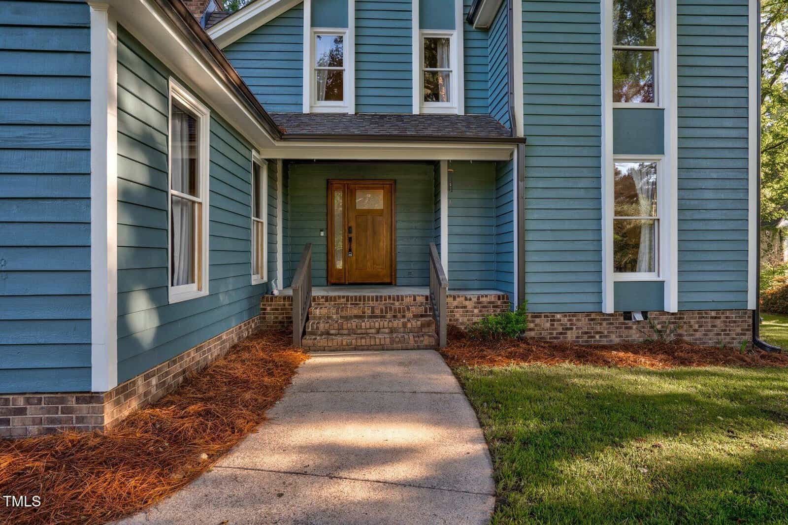 A front entryway of a blue house with vertical siding, brick foundation, a wooden door with side windows, concrete walkway, small steps, and landscaped mulch along the base.