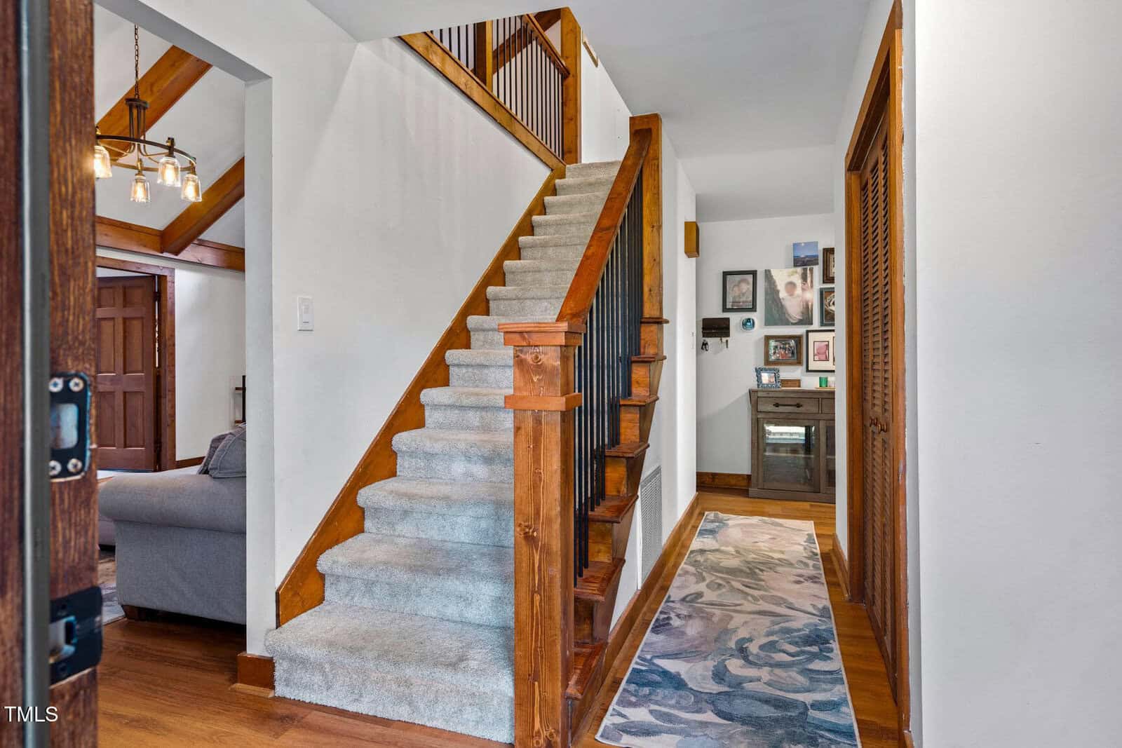 A carpeted staircase with wooden rails leads up from a hallway with hardwood floors. A patterned runner rug and framed pictures decorate the area. A glimpse of a living room with a sofa is visible to the left.