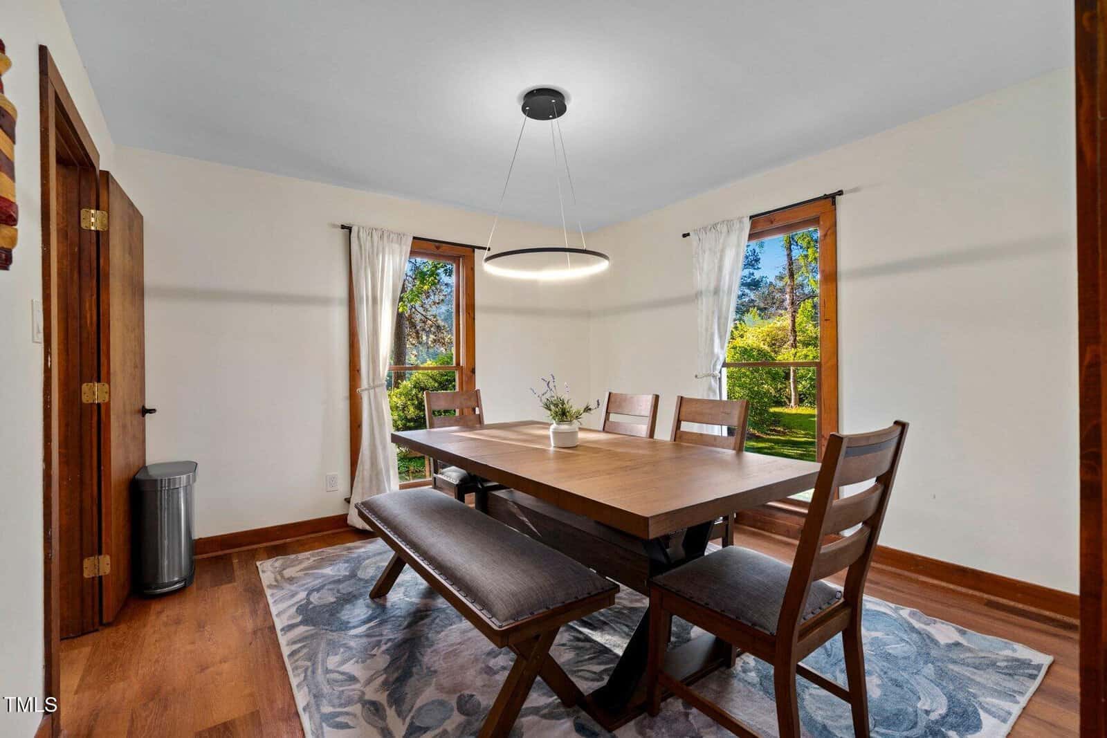 A dining room with a wooden table, six chairs, a bench, a modern circular ceiling light, two windows with white curtains, and a patterned rug on a wooden floor. Sunlight comes through the windows.