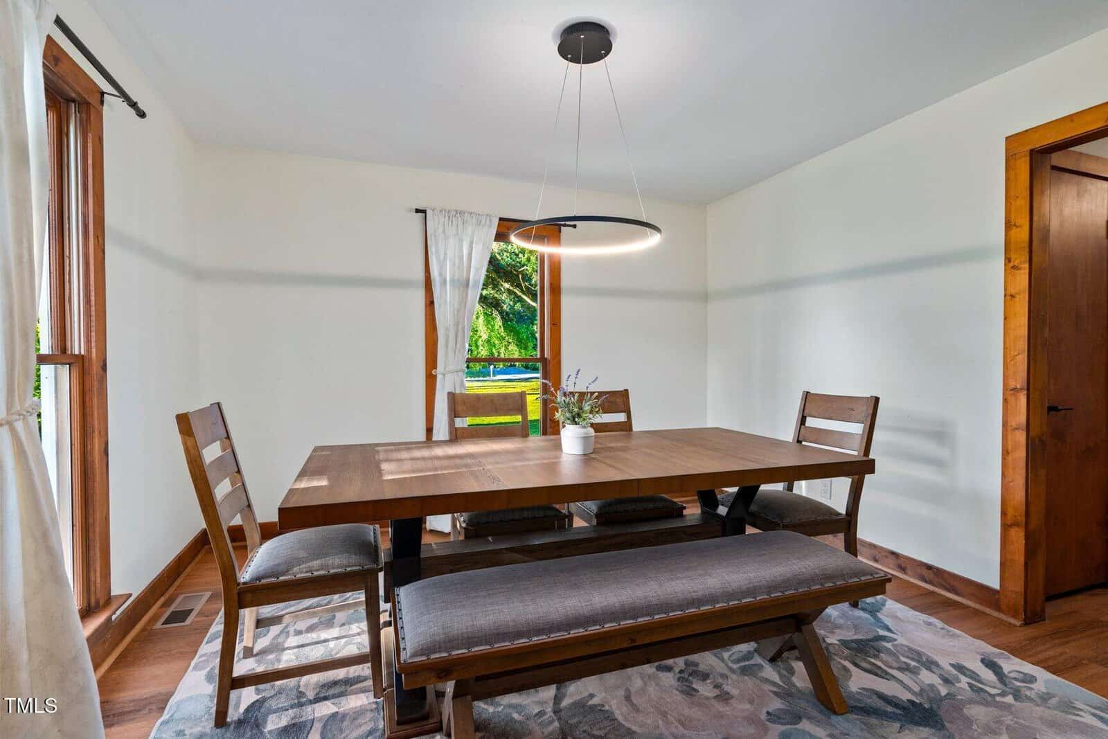 Dining room with a rectangular wooden table, three chairs, and a bench on a patterned rug. A modern circular light fixture hangs above. Windows with white curtains let in natural light. A small plant is on the table.