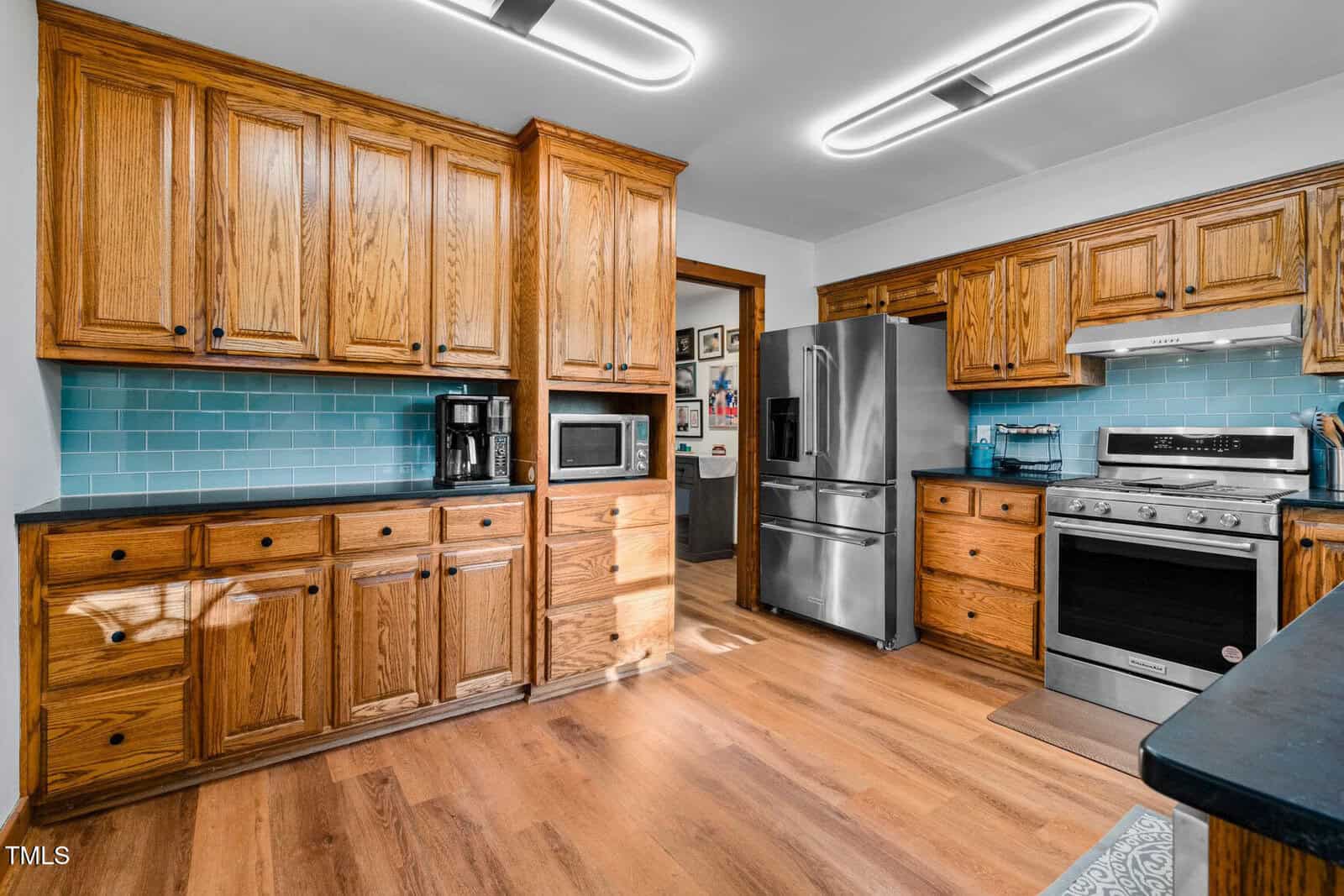 A kitchen with wooden cabinets, light blue subway tile backsplash, stainless steel refrigerator, stove, microwave, coffee maker, and wood flooring. Ceiling lights provide bright illumination.
