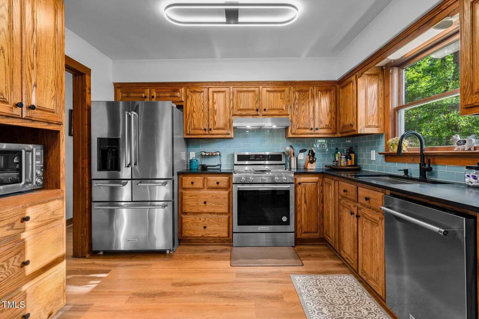 Modern kitchen with wooden cabinets, stainless steel appliances, blue tile backsplash, and large window above the sink. Hardwood floors and under-cabinet lighting are visible. Countertops hold various kitchen items.