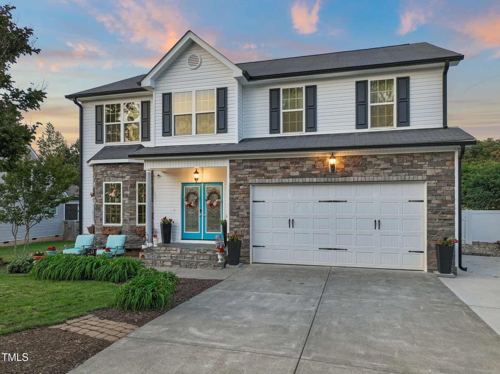 A two-story house with stone and white siding exterior, black shutters, and a double garage. The front yard has a green lawn, shrubs, and four teal chairs arranged near the entrance. The sky is partly cloudy at sunset.