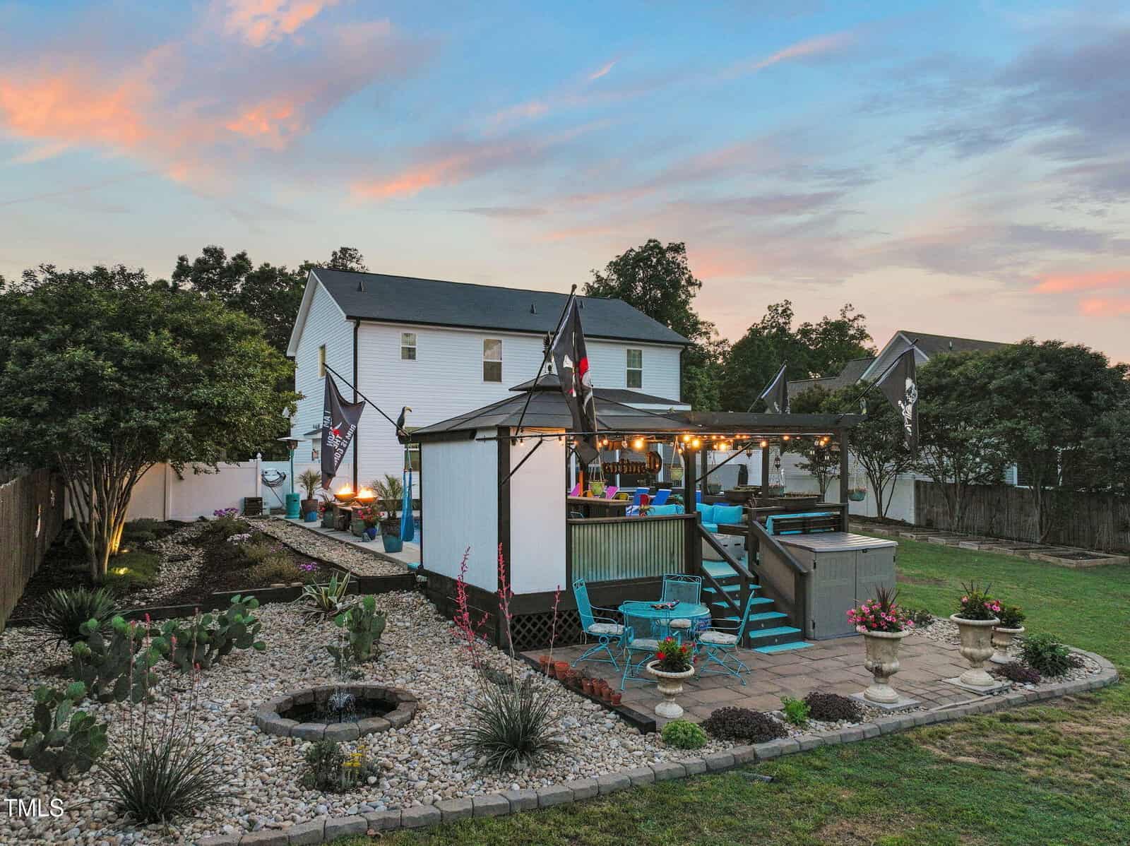 A backyard with a landscaped rock garden, small pond, and potted plants. There is a raised deck with seating, string lights, and flags. A white two-story house is in the background, surrounded by a fenced yard and trees.