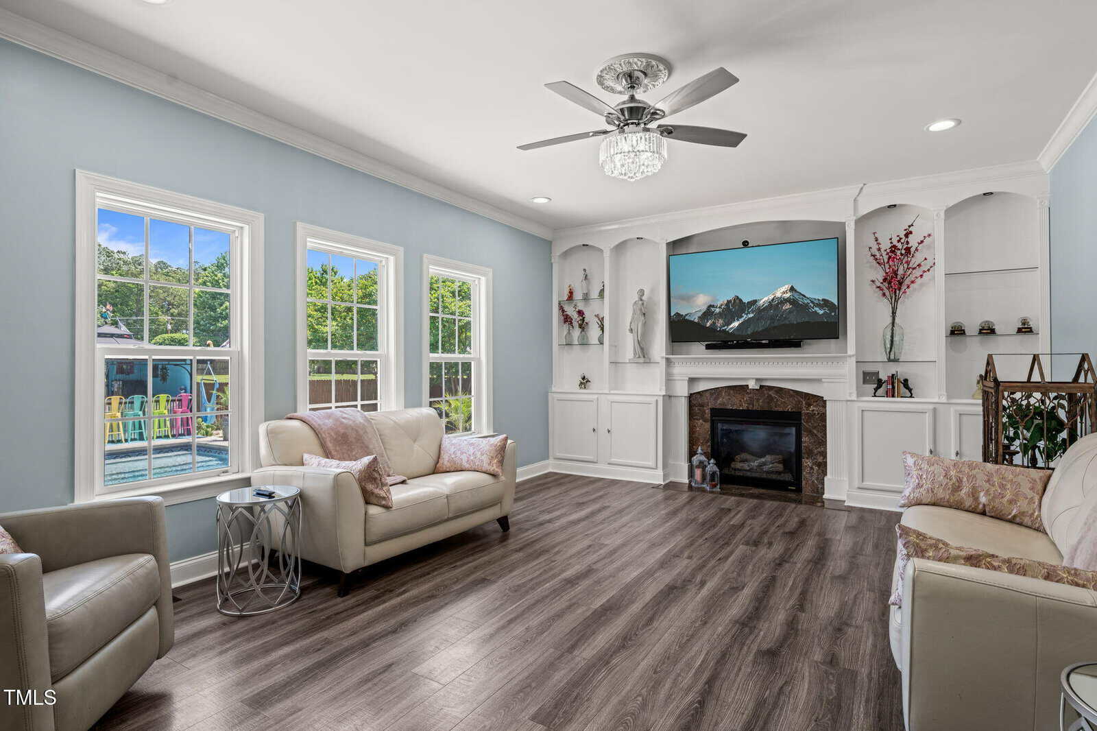 Living room with light blue walls, two cream sofas, a ceiling fan, wood flooring, built-in shelves, a fireplace, a TV displaying mountains, and large windows showing a backyard playground and trees.