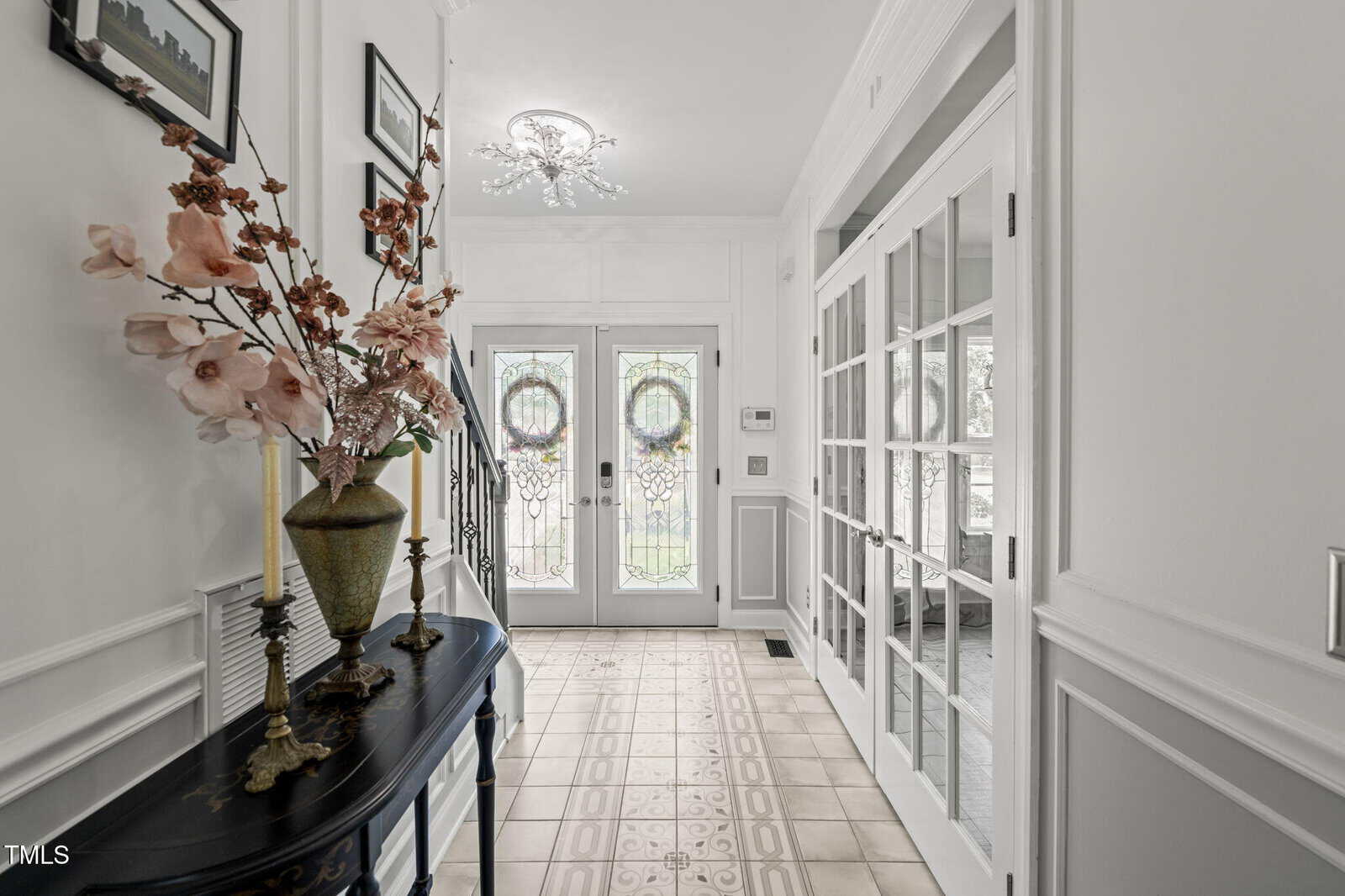 A bright hallway with white walls, decorative molding, and tiled flooring. A black console table with a floral vase and candles is on the left. Glass double doors with wreaths are at the far end, and French doors are on the right.