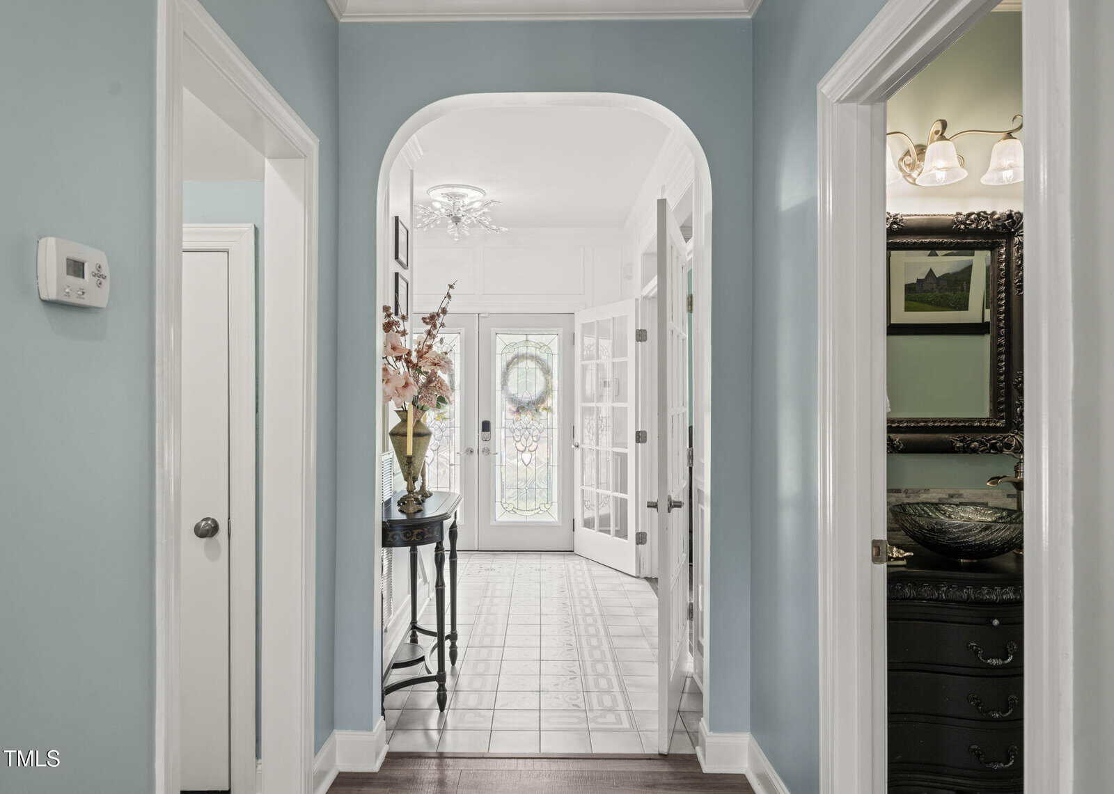 A hallway with light blue walls leads to a bright foyer with glass double doors. A small table with a vase of flowers is on the left. A bathroom with a sink and mirror is visible through an open door on the right.