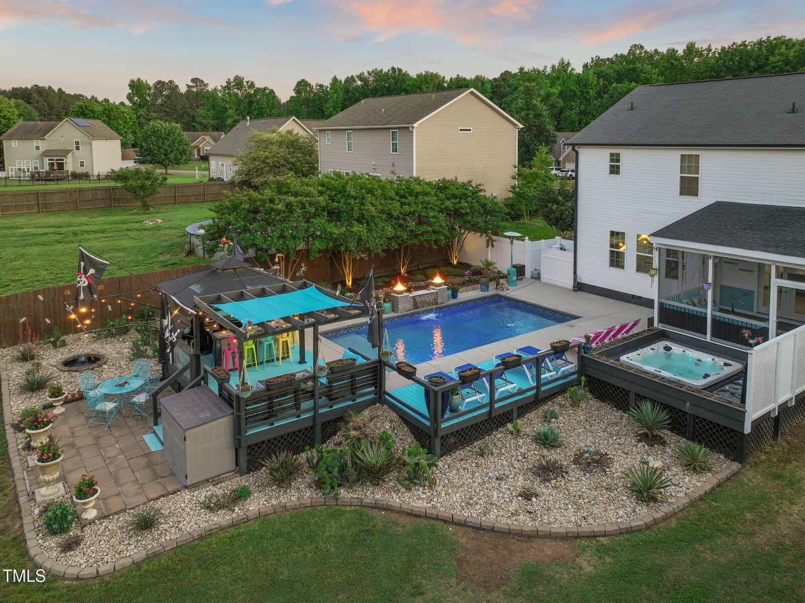 A backyard with a swimming pool, hot tub, and covered bar area with colorful stools. Several lounge chairs, string lights, a patio table, and landscaped garden beds surround the pool. Nearby houses and trees are visible.
