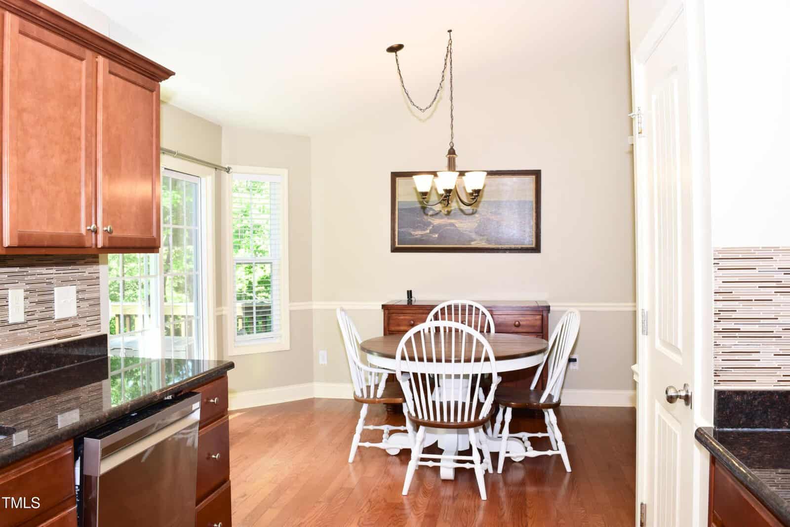 A bright kitchen dining area with a round wooden table, four white chairs, a sideboard, wood floors, and a window overlooking greenery. A chandelier hangs above the table and a framed picture is on the wall.