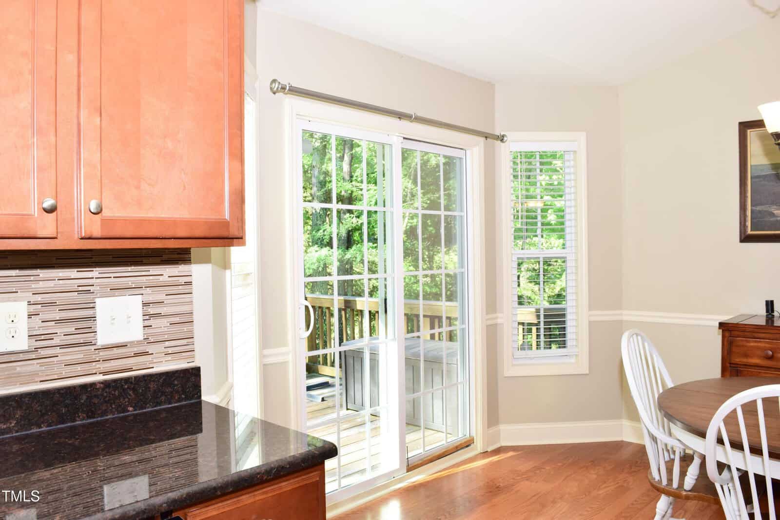 A kitchen with wood cabinets, black countertops, and a tile backsplash. A sliding glass door opens to a deck outside. A round wooden table with white chairs sits near a window with white blinds.
