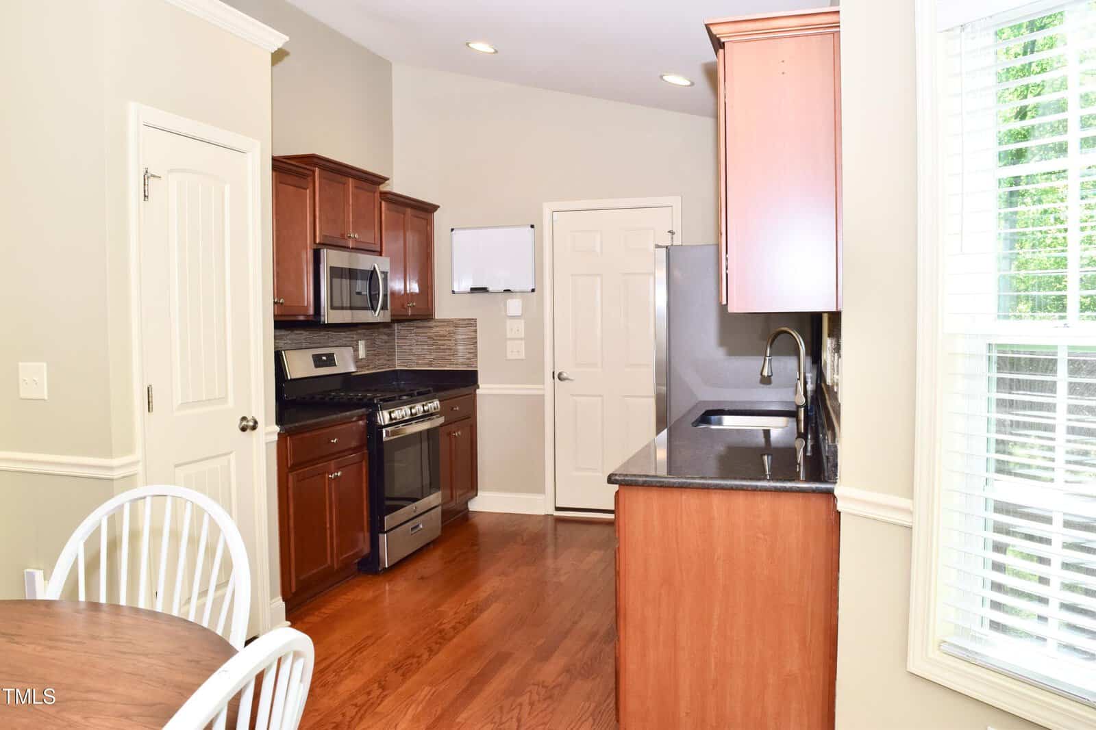 A kitchen with wood cabinets, stainless steel appliances, dark countertops, and a tile backsplash. A round dining table with white chairs is in the foreground near a window with white blinds.