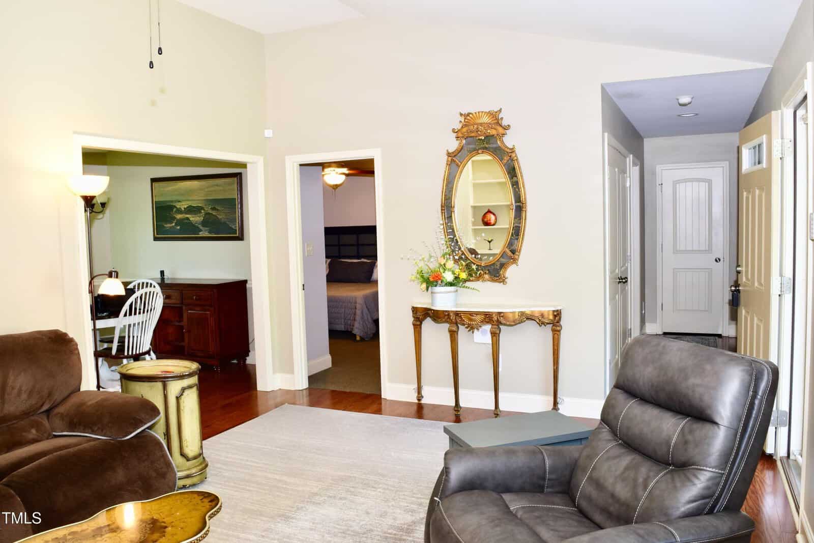 A living room with tan walls, leather recliners, a side table, and a console table topped with a flower arrangement beneath an ornate mirror. Doorways lead to a dining area, bedroom, and hallway.