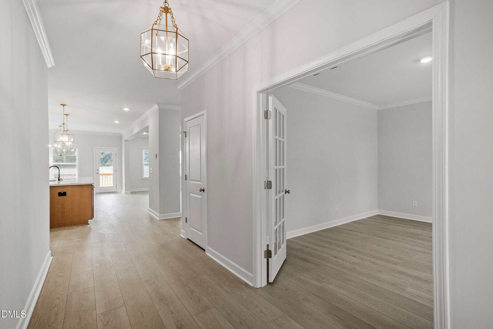 Bright hallway with light wood floors, neutral walls, and pendant lights. Glass French doors open to a room on the right; the hallway leads to a kitchen and living area with large windows in the background.