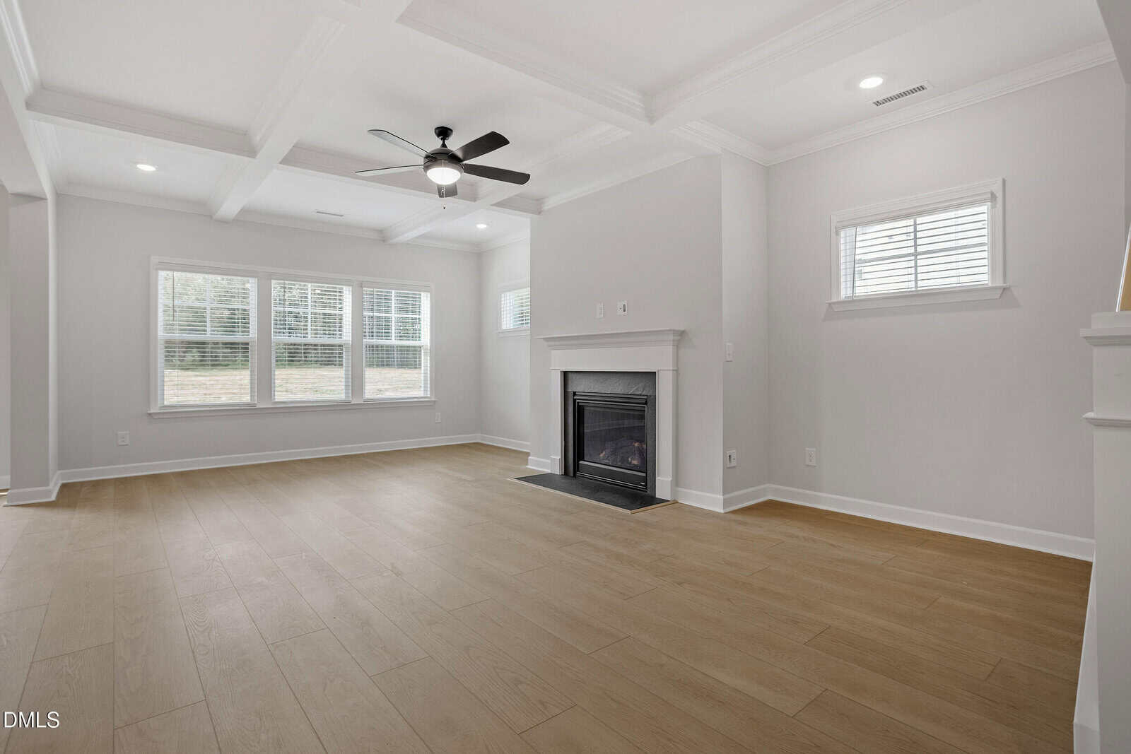 A spacious, empty living room with light wood floors, white walls, a ceiling fan, recessed lighting, multiple windows, and a white fireplace. Natural light fills the room through the large windows.