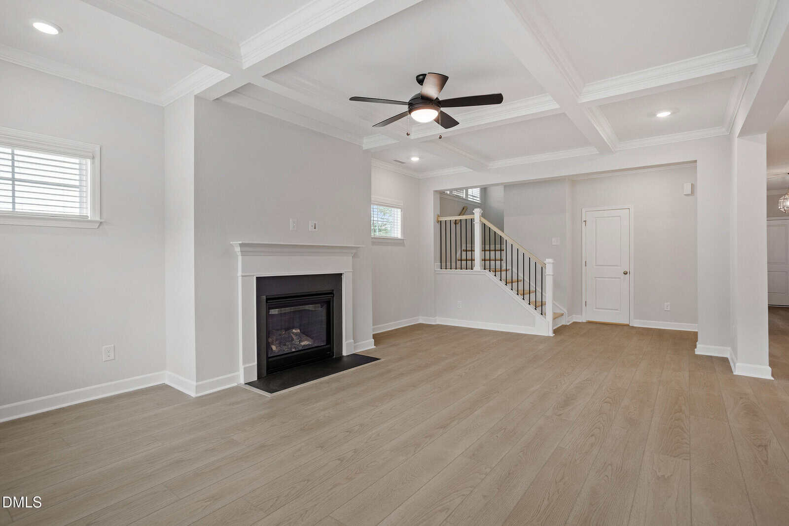 Bright living room with light wood floors, a white coffered ceiling, black ceiling fan, fireplace, two windows, and a staircase with wooden handrail leading to the upper floor. White walls and trim throughout.