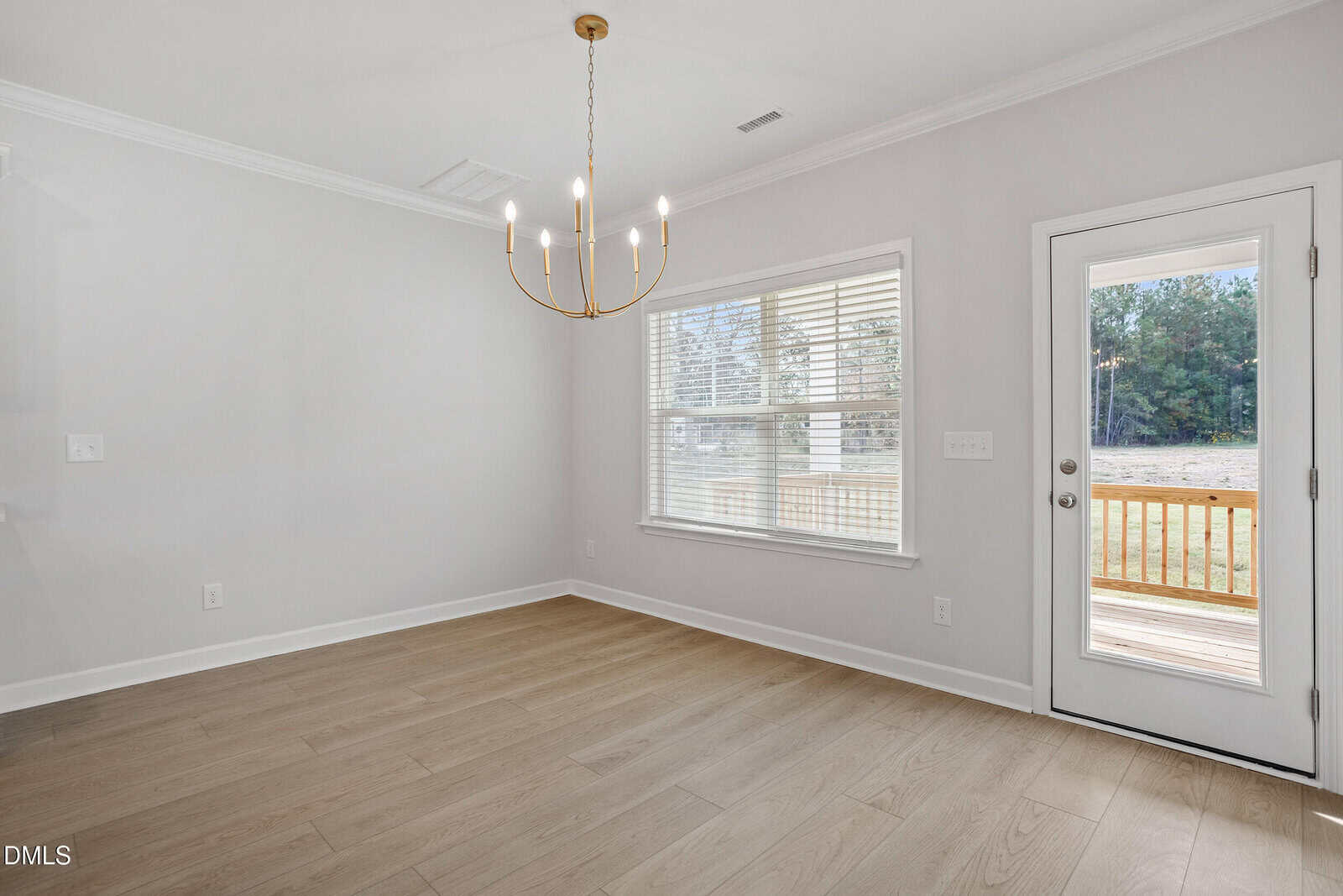 A bright, empty room with light wood floors, white walls, a window with blinds, a modern gold chandelier, and a door leading to a wooden deck outside.