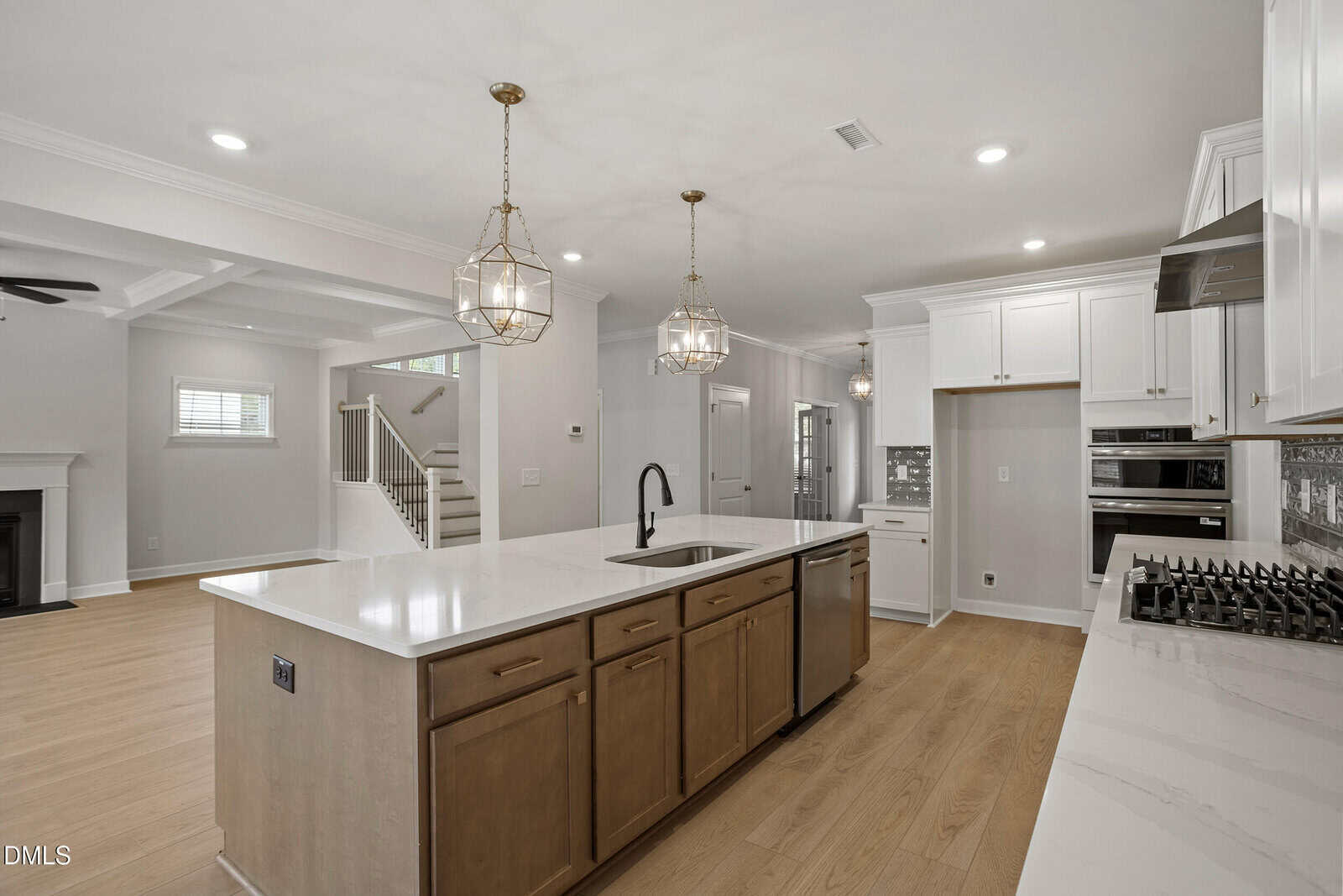 Modern kitchen with a large island featuring a sink and dishwasher, light wood flooring, white cabinets, stainless steel appliances, and two geometric pendant lights. Open layout leads to stairs and a living area with a fireplace.
