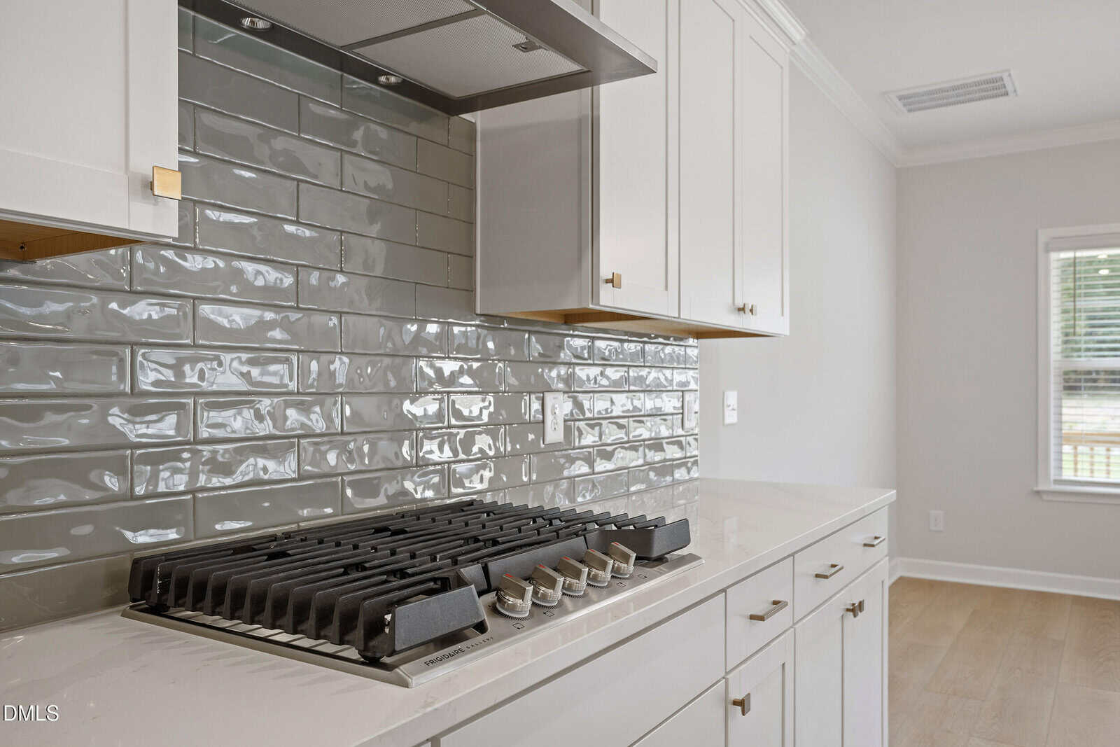 A modern kitchen with a stainless steel gas cooktop, white cabinets, and a glossy, gray subway tile backsplash reflecting light. Light wood flooring and a window with blinds are also visible.