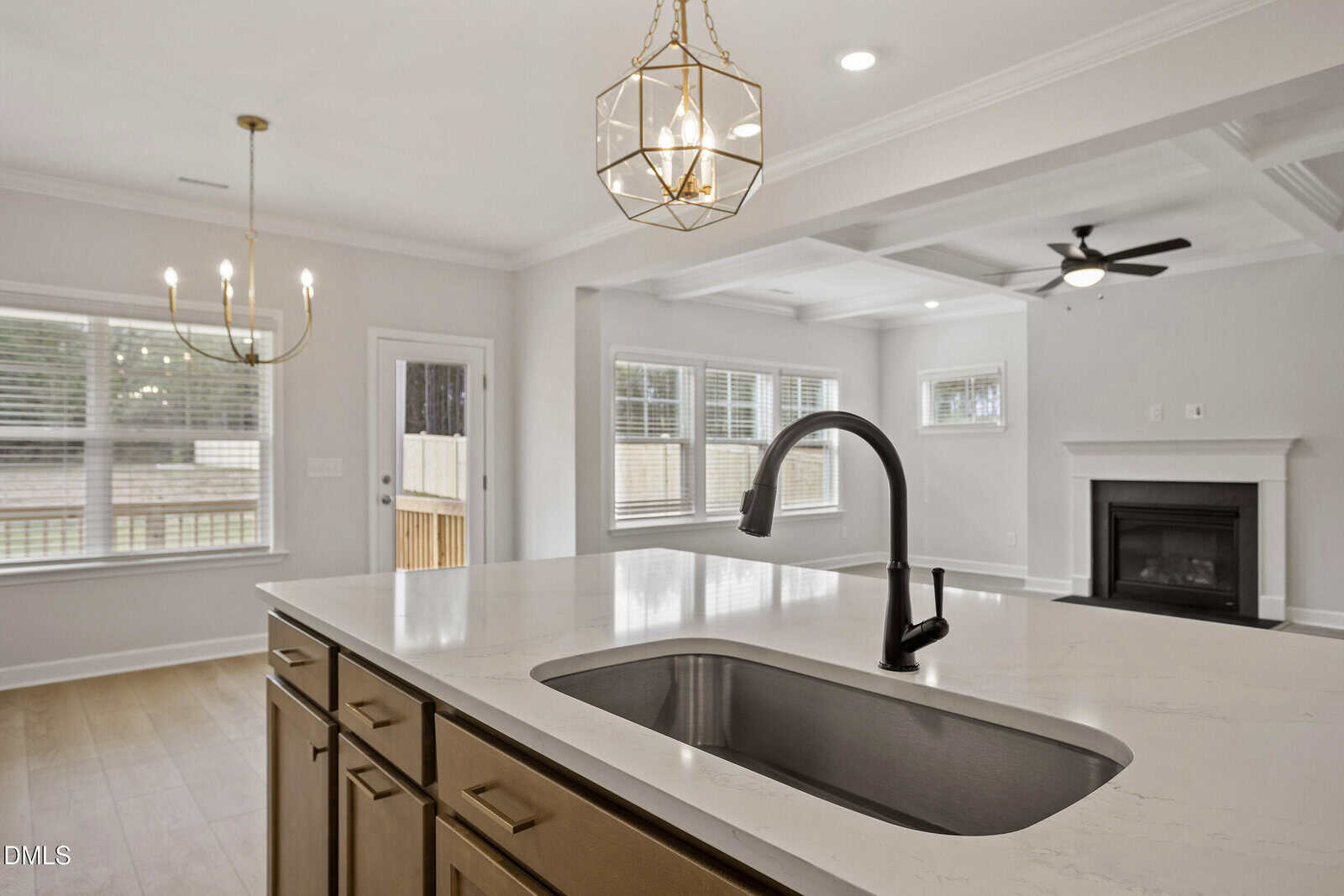 A modern kitchen with a marble island and sink in the foreground, pendant lighting above, and an open view into a living area with a ceiling fan, fireplace, and large windows.