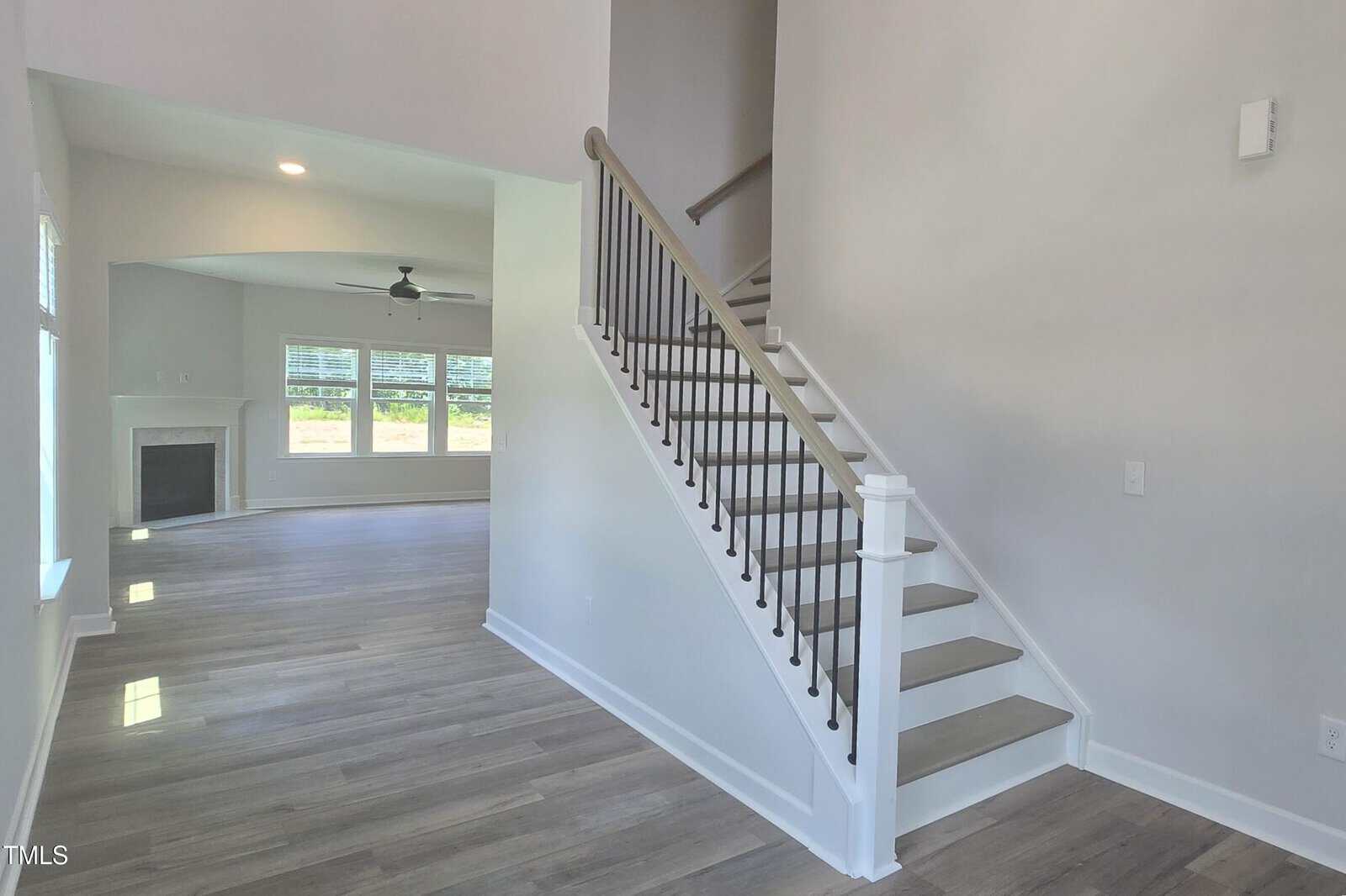 A modern home interior with light gray walls, wood flooring, a staircase with white steps and black railings, and a living area featuring large windows and a fireplace.