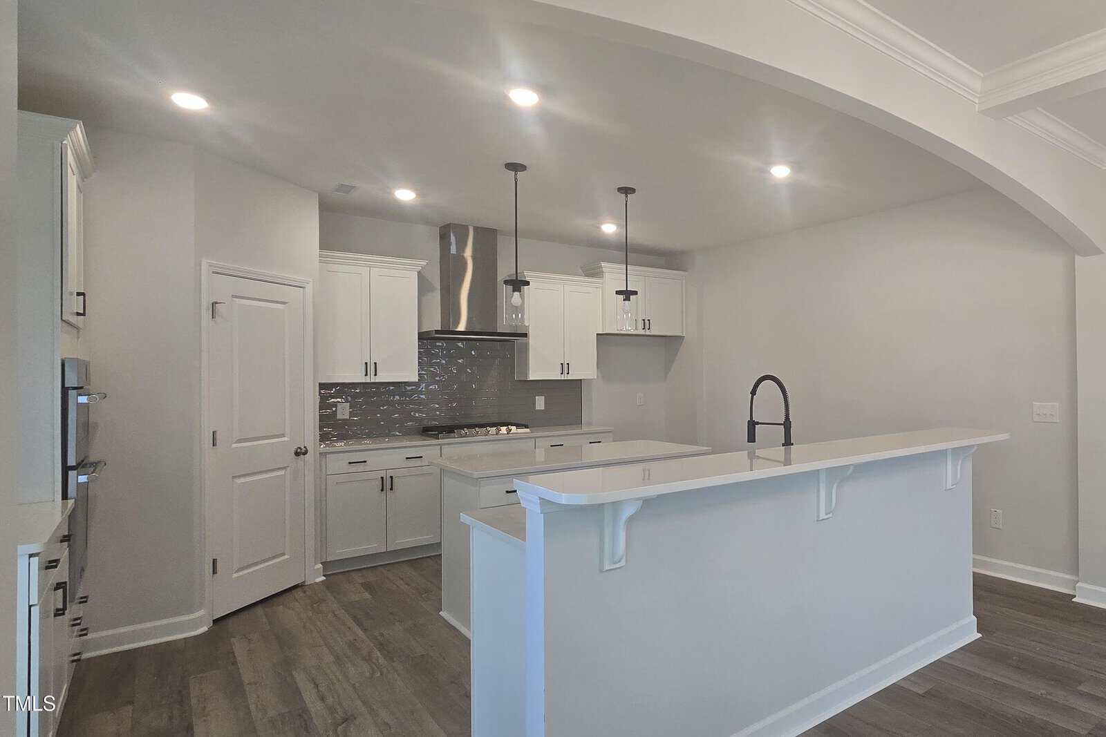 Modern kitchen with white cabinets, stainless steel appliances, tile backsplash, large island with sink and high-arc faucet, pendant lights, wood flooring, and recessed ceiling lights. Walls and trim are painted light neutral colors.