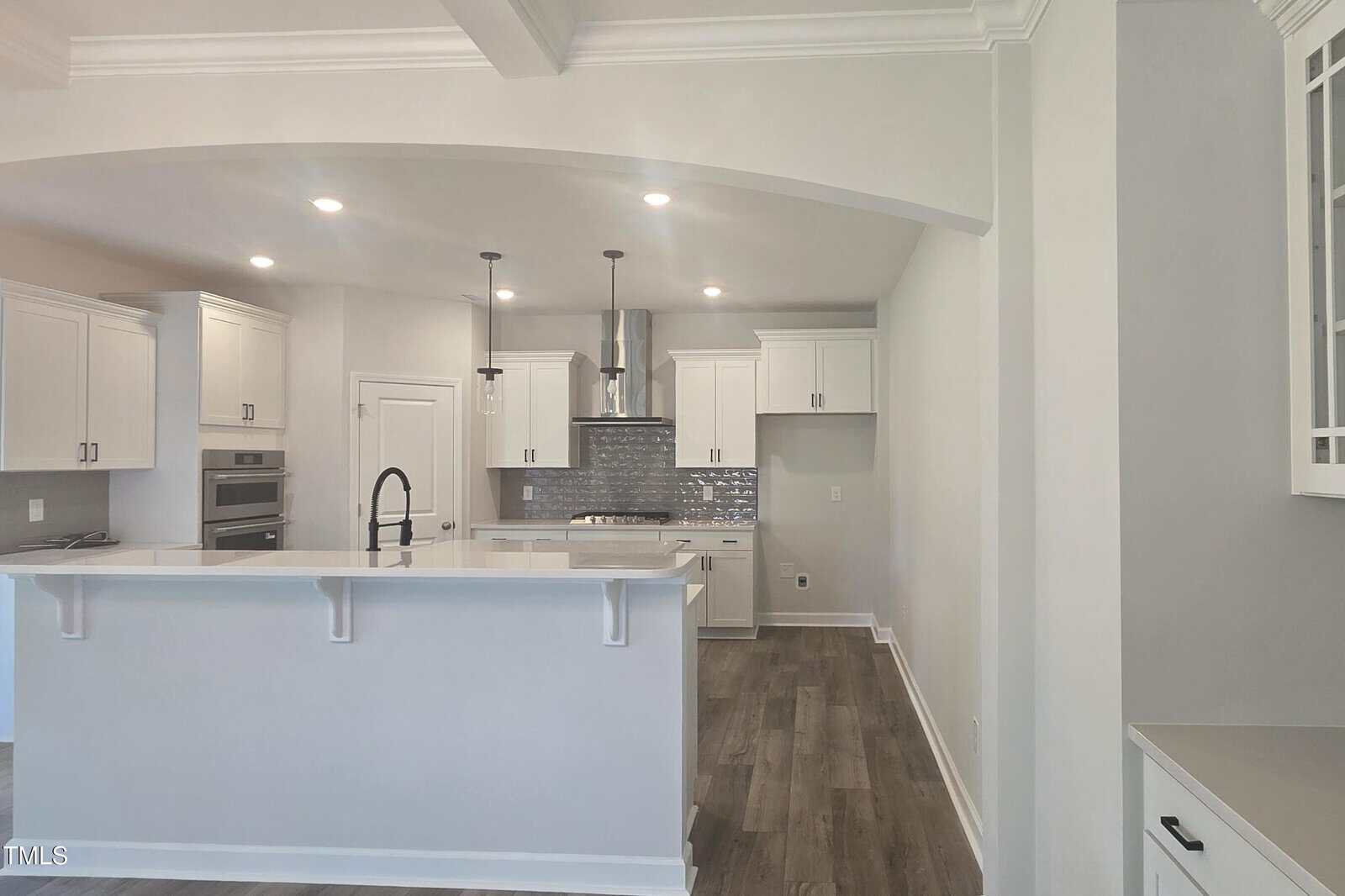 Modern kitchen with white cabinets, a large island with sink and faucet, stainless steel appliances, gray tile backsplash, pendant lights, and wood-look flooring. Walls and ceiling are painted white, creating a bright space.