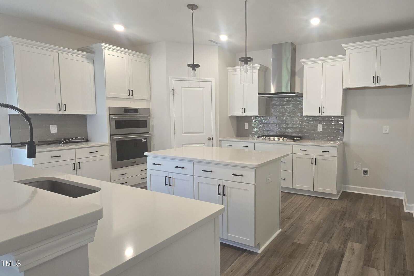 Modern kitchen with white cabinets, stainless steel appliances, a central island, pendant lights, gray tile backsplash, wood-look flooring, and ample counter space. The overall design is bright and spacious.