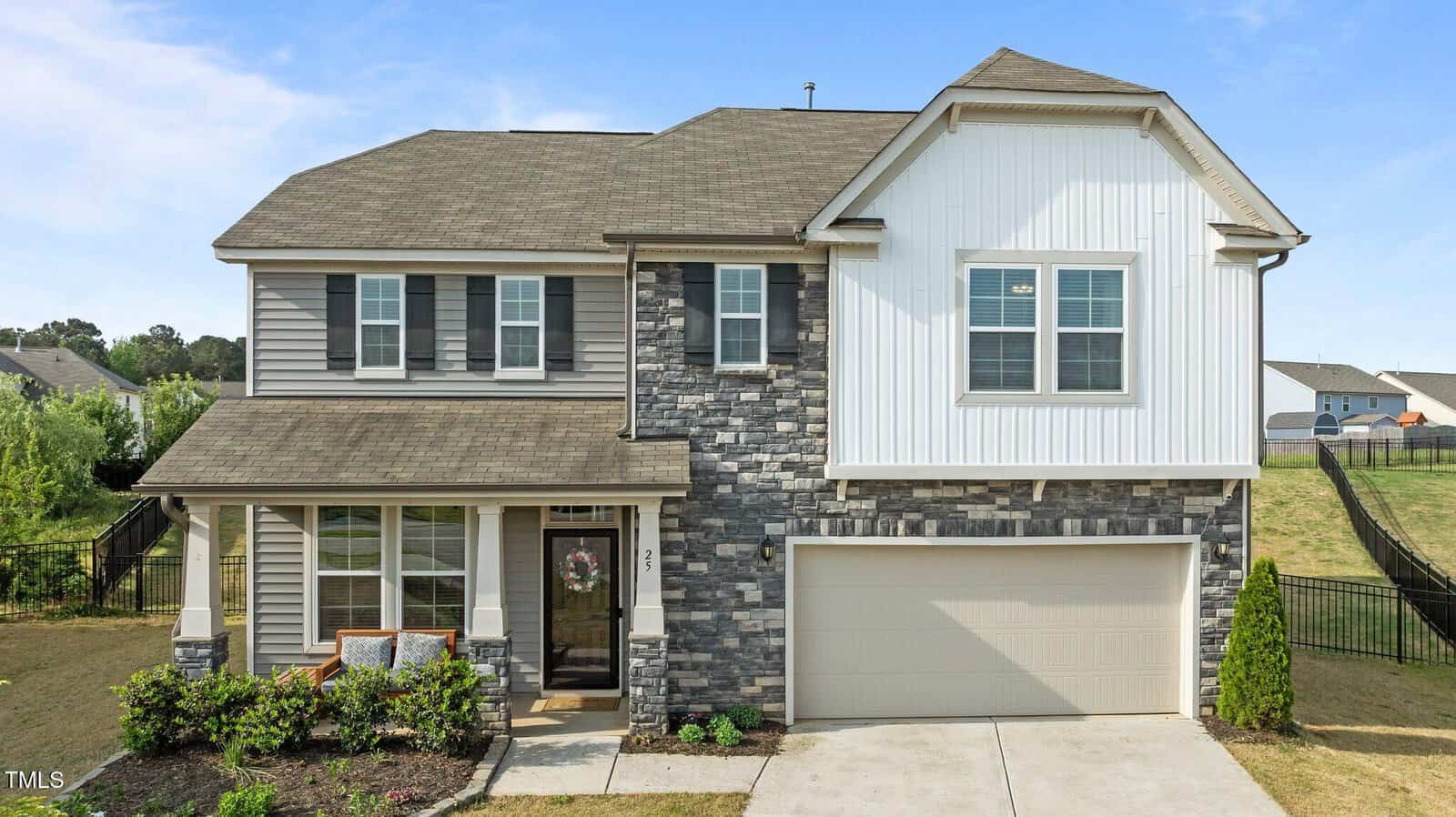 A modern two-story house with a mix of gray stone and white siding, a covered front porch with a wreath on the door, two-car garage, and a neatly landscaped front yard under a blue sky.