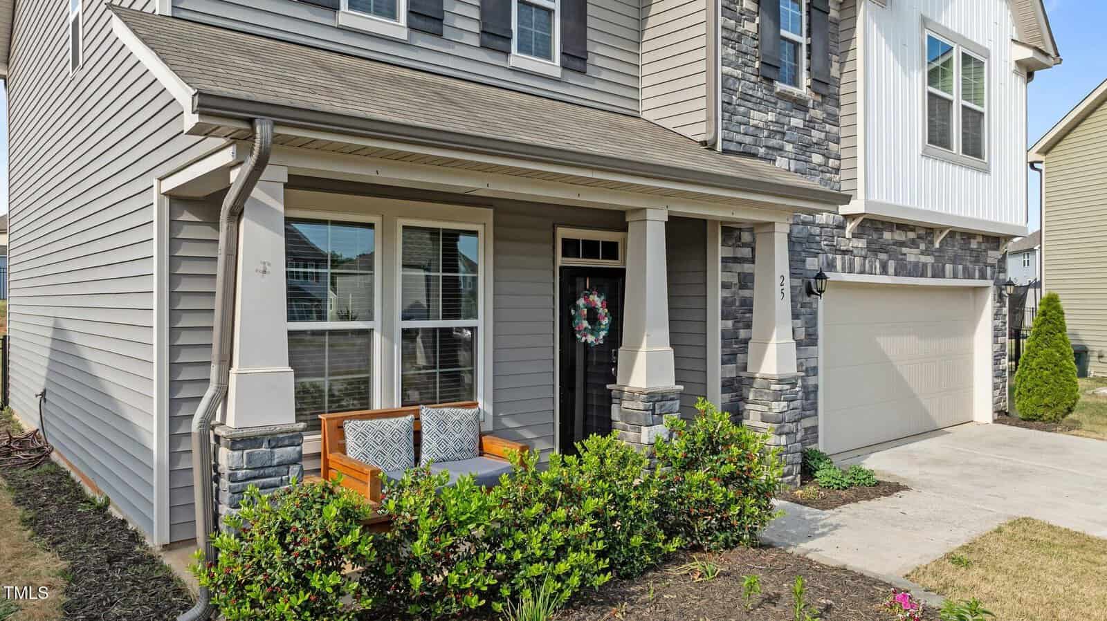 A two-story house with gray siding and stone accents, a covered front porch with a wooden bench and cushion, and a decorated front door. A driveway leads to an attached garage; shrubs line the porch.