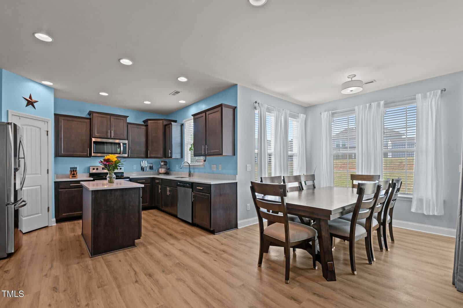 Modern kitchen and dining area with wooden floors, dark brown cabinets, light blue walls, stainless steel appliances, a rectangular dining table with eight chairs, and large windows with white curtains letting in natural light.