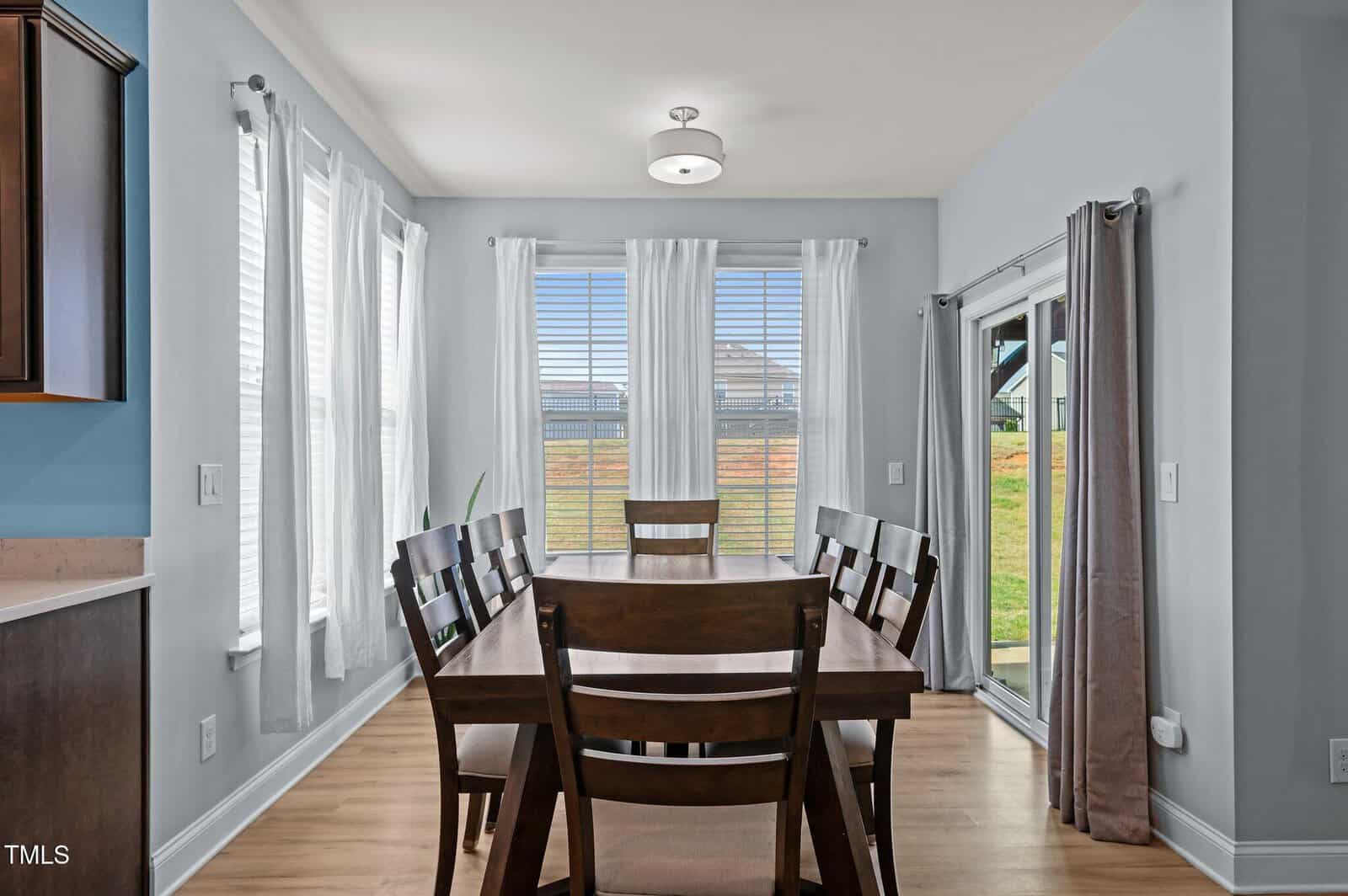A dining area with a wooden table and six chairs, surrounded by large windows with white curtains. Outside, grass and a hill are visible. Sliding glass doors are on the right, and light streams into the room.