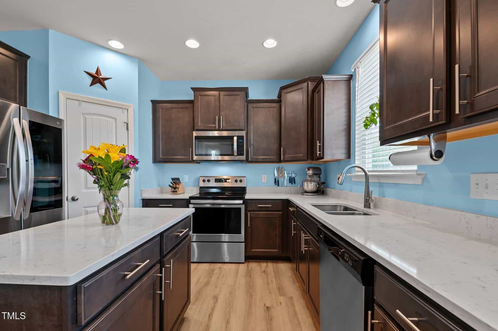 Modern kitchen with dark wood cabinets, stainless steel appliances, white quartz countertops, a vase of flowers on the island, blue walls, and wood flooring. A paper towel holder is by the sink under a window with blinds.
