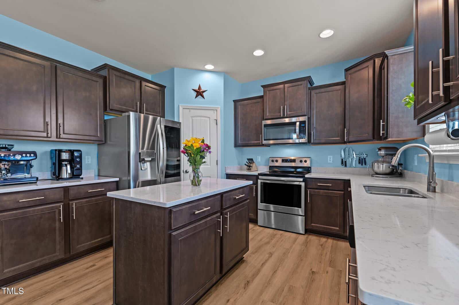 Modern kitchen with stainless steel appliances, dark wood cabinets, light countertops, and a center island with a vase of colorful flowers. The walls are painted light blue and there is a star decoration above the door.
